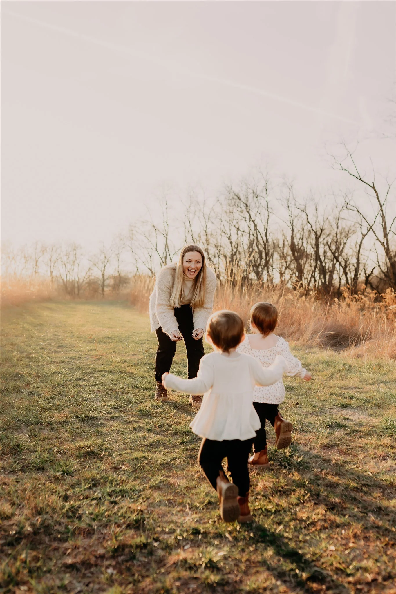 A woman and two young children running and playing outdoors on a grassy field during sunset during sunset in autumn at Columbia, MO brand photography session at Rock Bridge Memorial State Park.