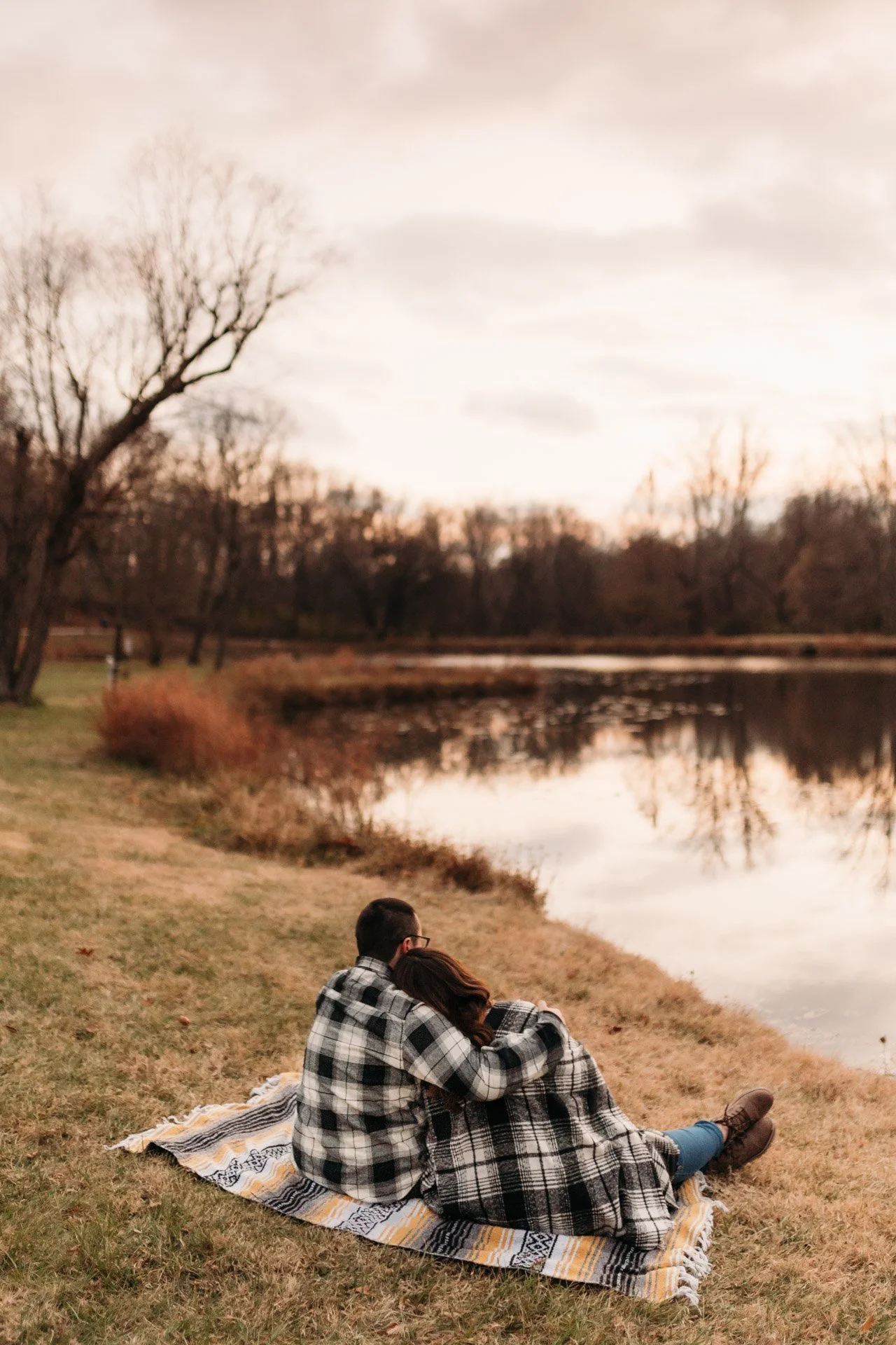 A couple sits on a blanket by a lake, embracing in a park during sunset with leafless trees in the background at their engagement photography session at Queeny Park in STL.