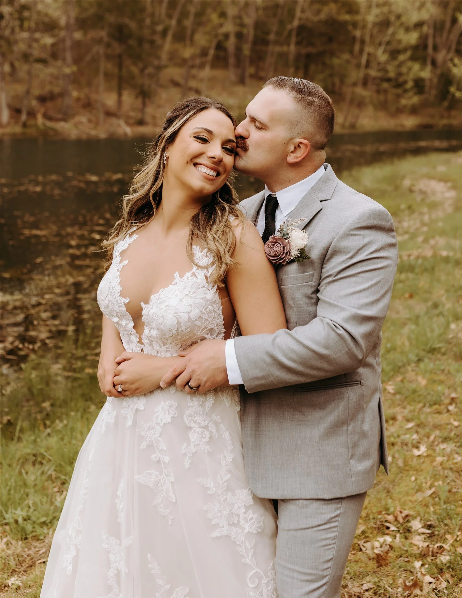 A happy couple in wedding attire standing outdoors near a pond, with trees in the background at Rolla, MO rustic wedding venue, The Village. Photos by Columbia, MO Photographer Liv Strange Photography.