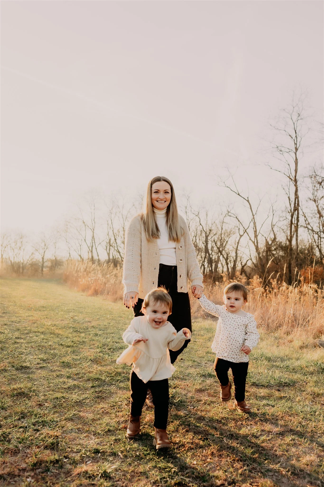 A woman and two young children running outdoors on a grassy field during sunset, with bare trees in the background during sunset in autumn at Columbia, MO brand photography session at Rock Bridge Memorial State Park.