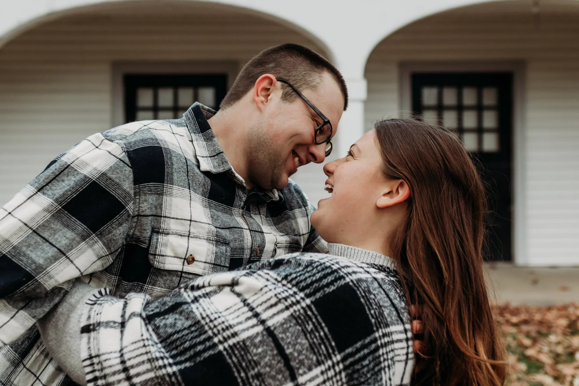A smiling couple in plaid shirts share a close moment outdoors in front of a house at their engagement photography session at Queeny Park in STL.