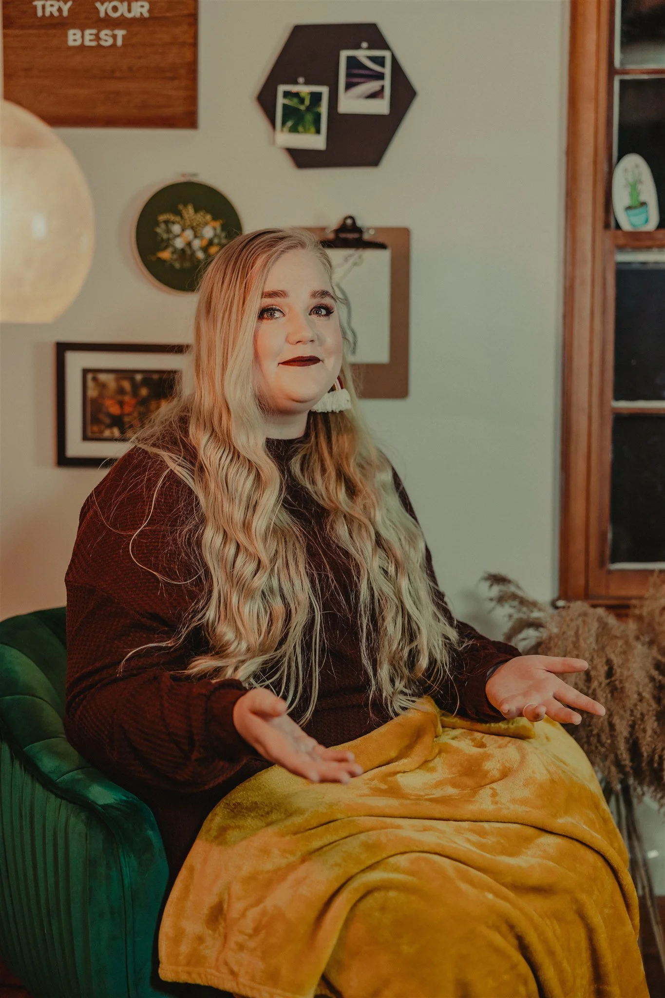 A woman with long, wavy blonde hair, wearing dark lipstick and a dark top, is sitting on a green velvet chair with a yellow blanket on her lap during a trauma informed photography session in Columbia, Missouri.