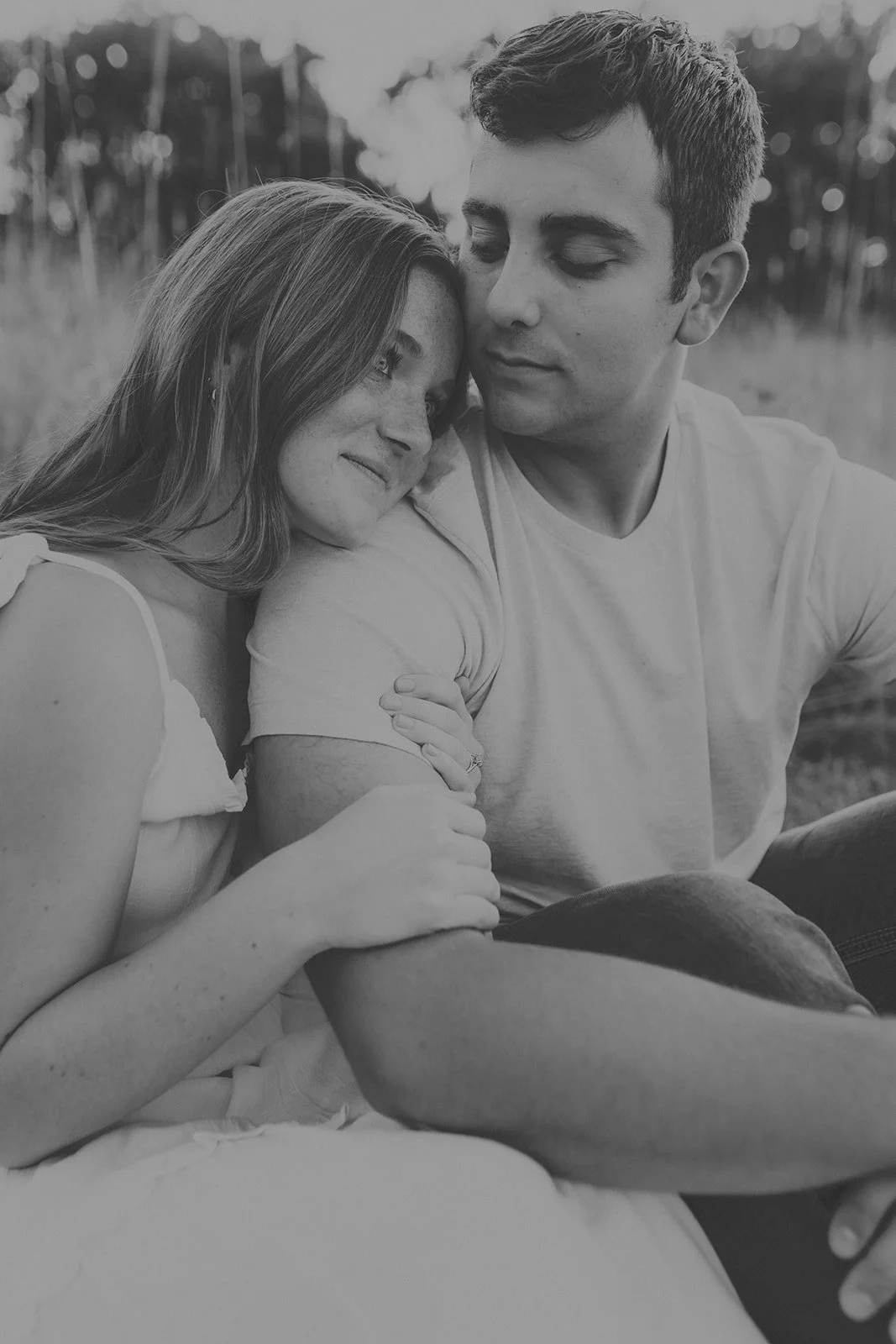 A black-and-white photo of a couple sitting closely together outdoors, with the woman resting her head on the man's shoulder and both looking peaceful and happy during their engagement photography session at Rock Bridge State Park in Columbia, MO.
