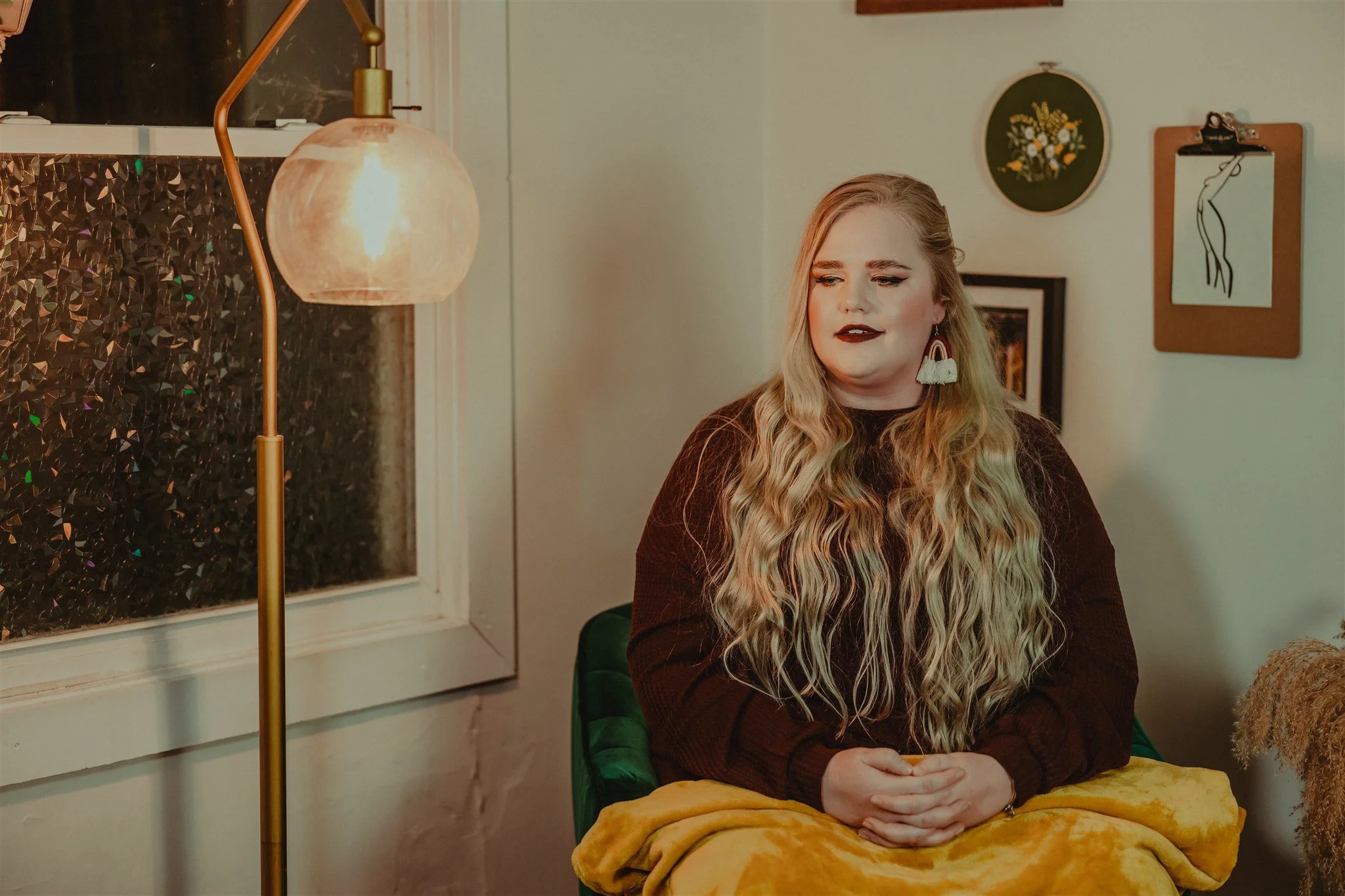 A woman with long, wavy blonde hair and dark lipstick sits on a green chair in a cozy, softly lit room. She is wearing a dark sweater, colorful earrings, and a yellow blanket over her lap during a trauma informed photography session in Columbia, MO.