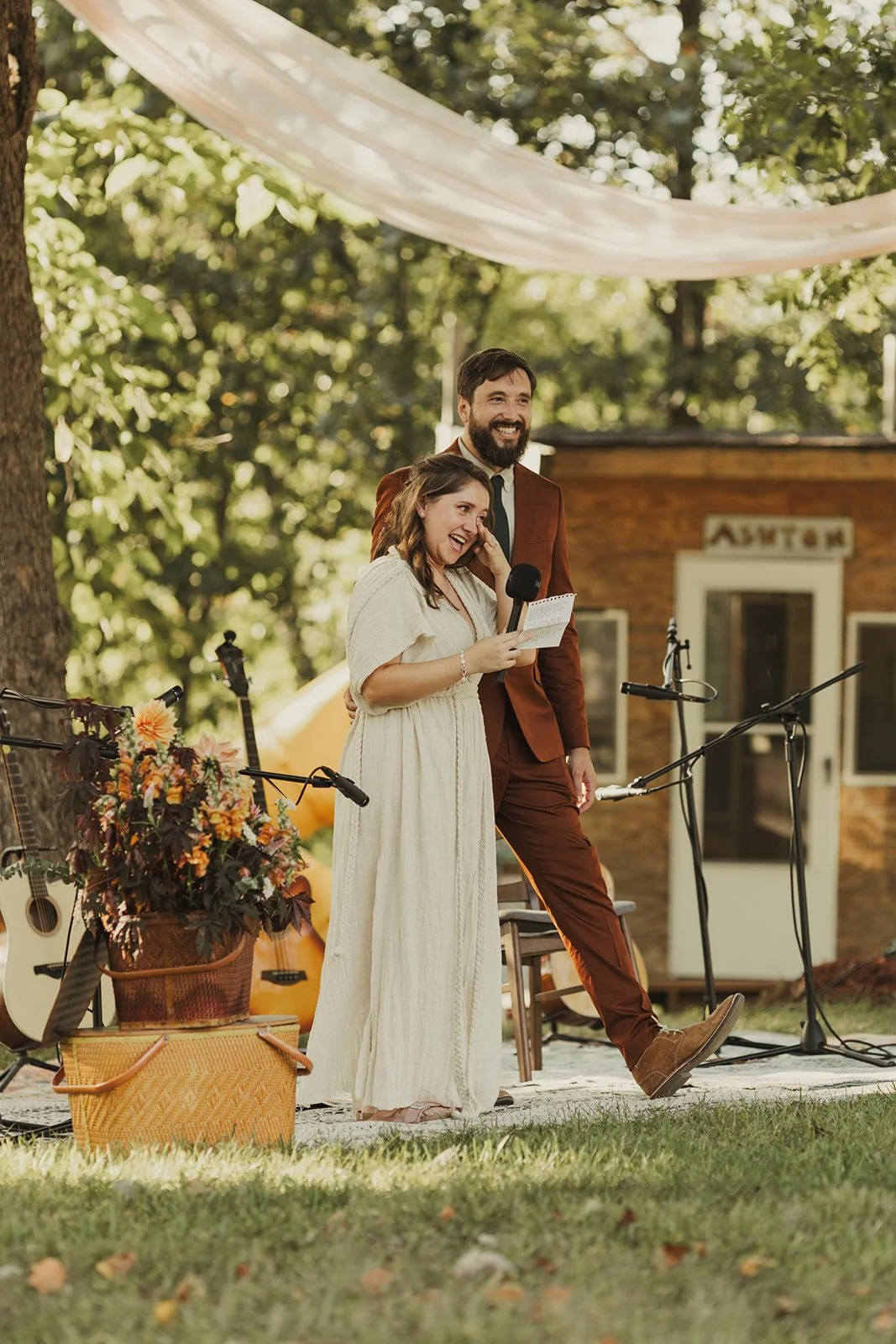A woman and man are standing together outdoors during daytime at a wedding, smiling and holding a microphone and a paper. The woman is wearing a cream-colored dress, and the man is wearing a brown suit. There are guitars and a basket of flowers nearby, with a small wooden building in the background.