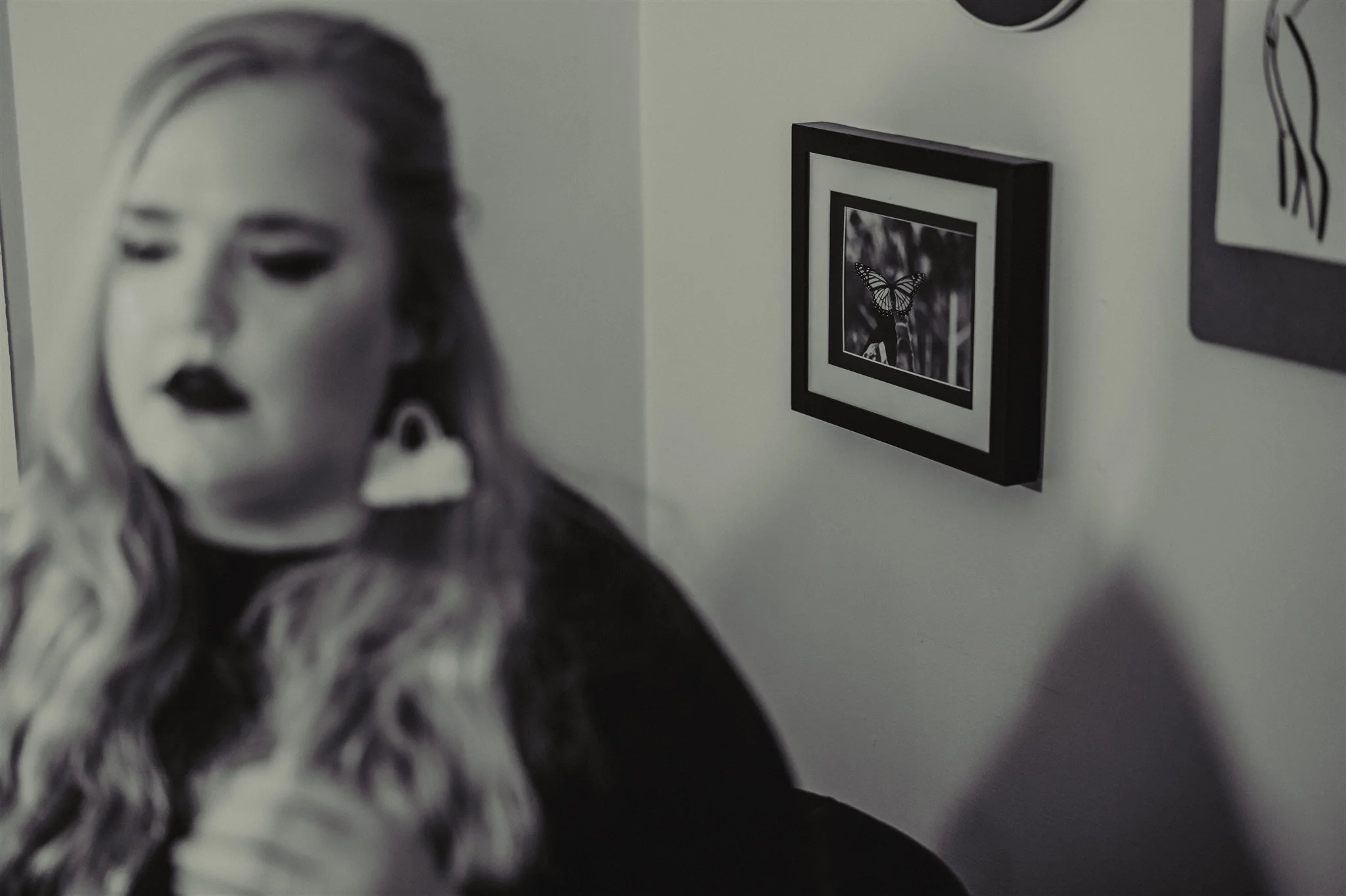 A blurred portrait of a woman with dark lipstick and blonde hair, and a framed picture of a butterfly on the wall in black and white during a trauma informed photography session in Columbia, Missouri.