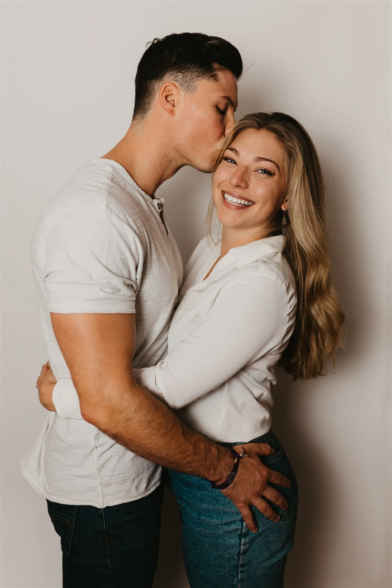 A man is kissing a smiling woman on the forehead as they embrace against a plain white wall.