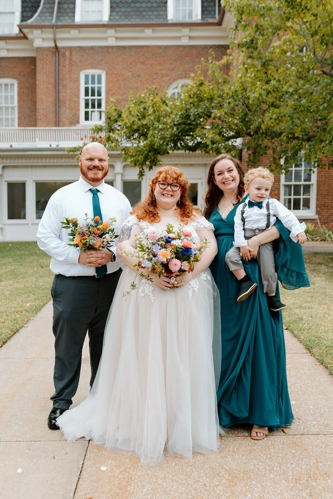 Group of four people, including a bride, standing outdoors in front of a large brick house, holding bouquets, with one woman carrying a young boy at an LGBTQ+ wedding in Columbia, MO at wedding venue, Stephens College Senior Hall.