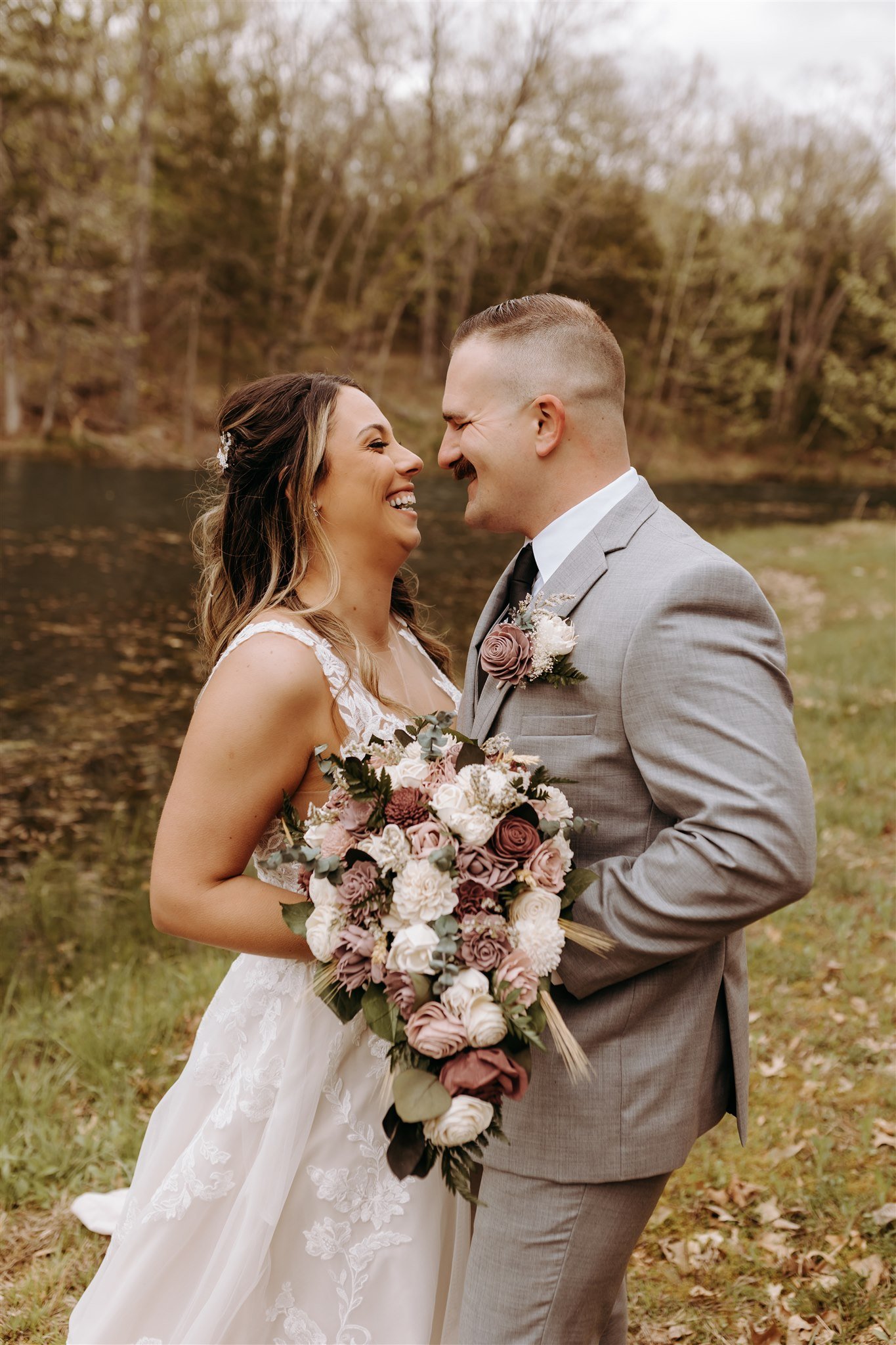 A bride and groom are smiling and leaning close to each other near a pond, with trees in the background at Rolla, MO rustic wedding venue, The Village. Photos by Columbia, MO Photographer Liv Strange Photography.