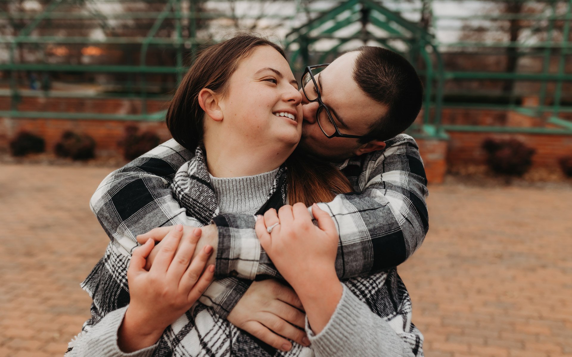 A couple in plaid shirts sharing a warm embrace and a kiss outdoors on a brick-paved area with green metal structures in the background at their engagement photography session at Queeny Park in STL.
