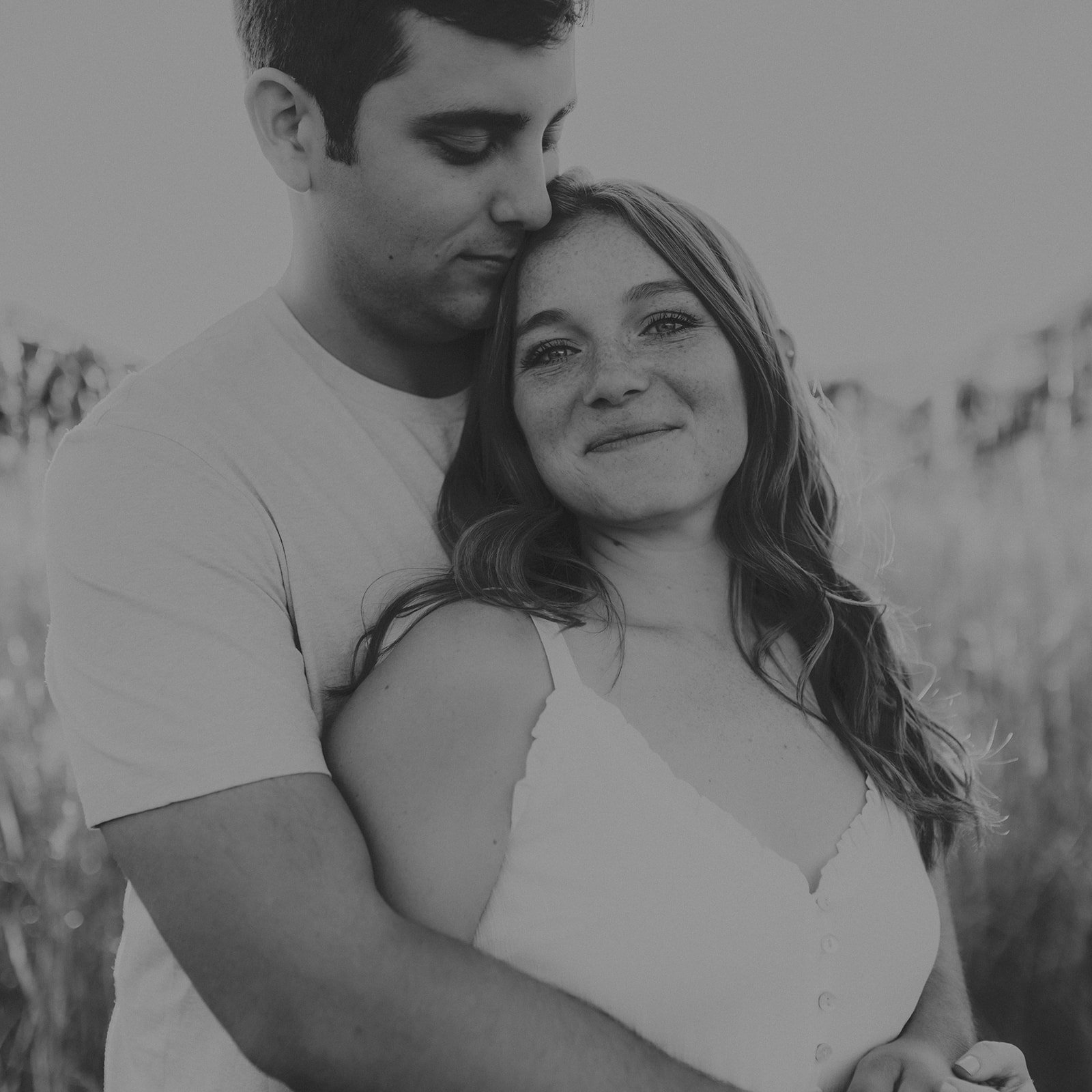 A black and white photo of a young man and woman embracing outdoors, with the man gently resting his head on the woman's head, both smiling softly during their engagement photography session at Rock Bridge State Park in Columbia, MO.