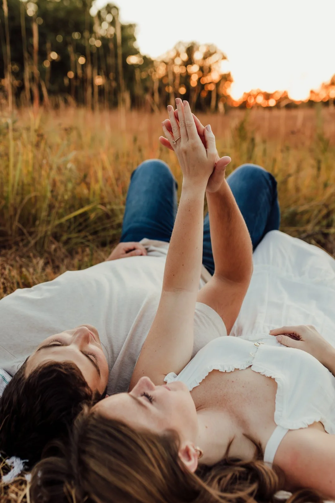 A young engaged couple lying in a field at sunset, holding hands and looking at each other during their couples photography session at Rock Bridge State Park in Columbia, MO.