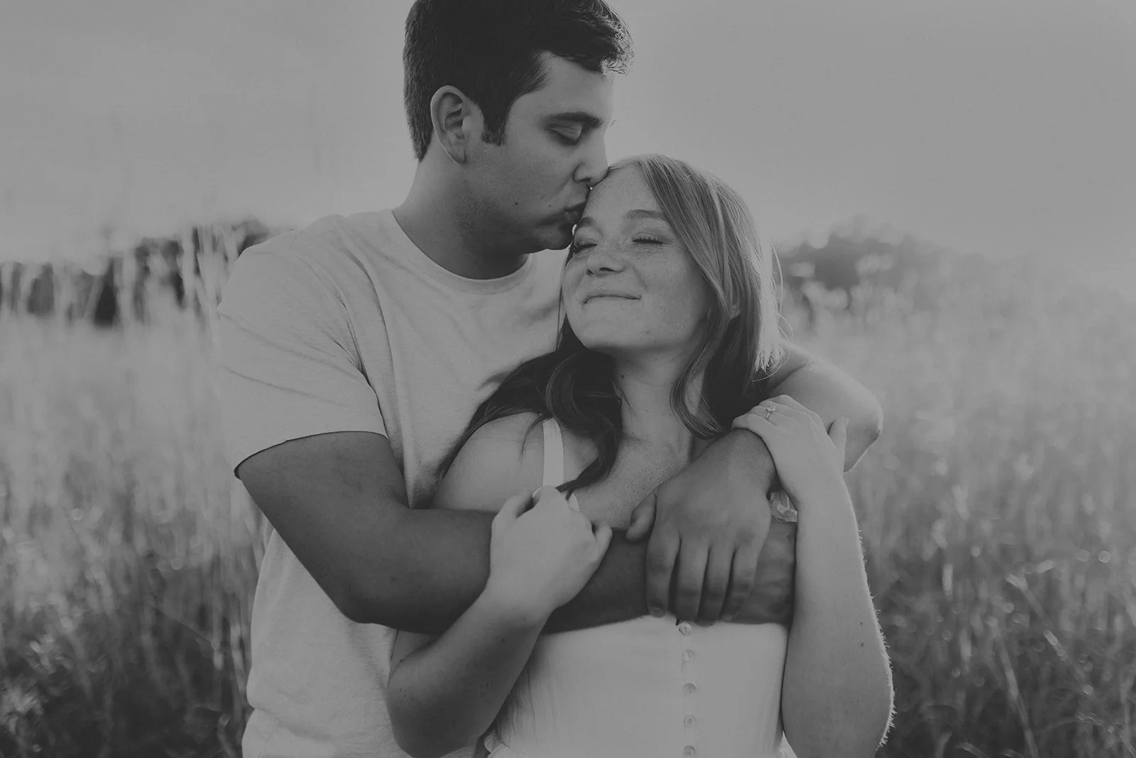 A black-and-white photo of a man kissing a woman on the forehead while hugging her, outdoors in a field during their engagement photography session at Rock Bridge State Park in Columbia, MO.