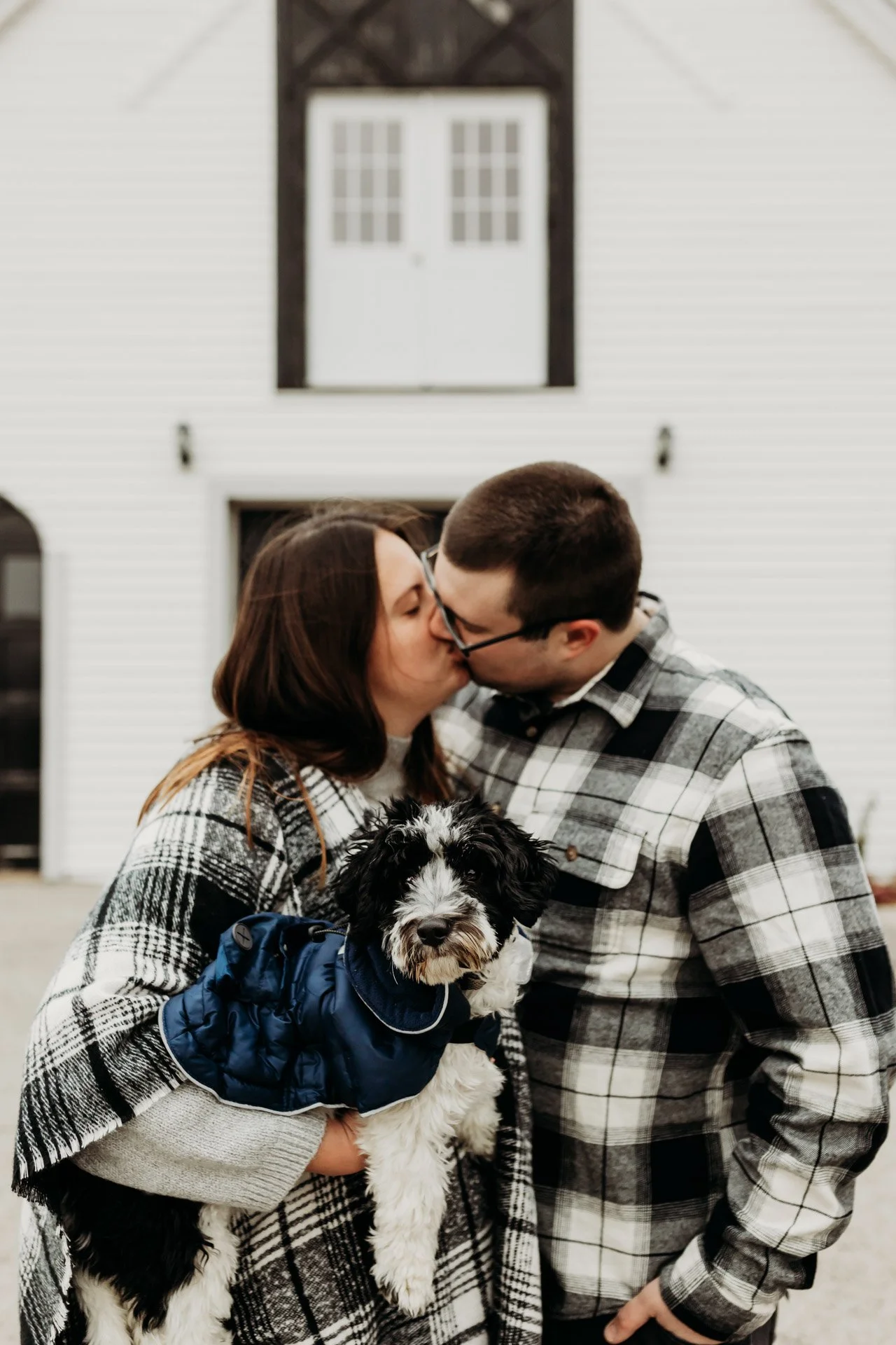 A couple kissing while holding a black and white puppy in front of a white house with a barn door balcony at their engagement photography session at Queeny Park in STL.