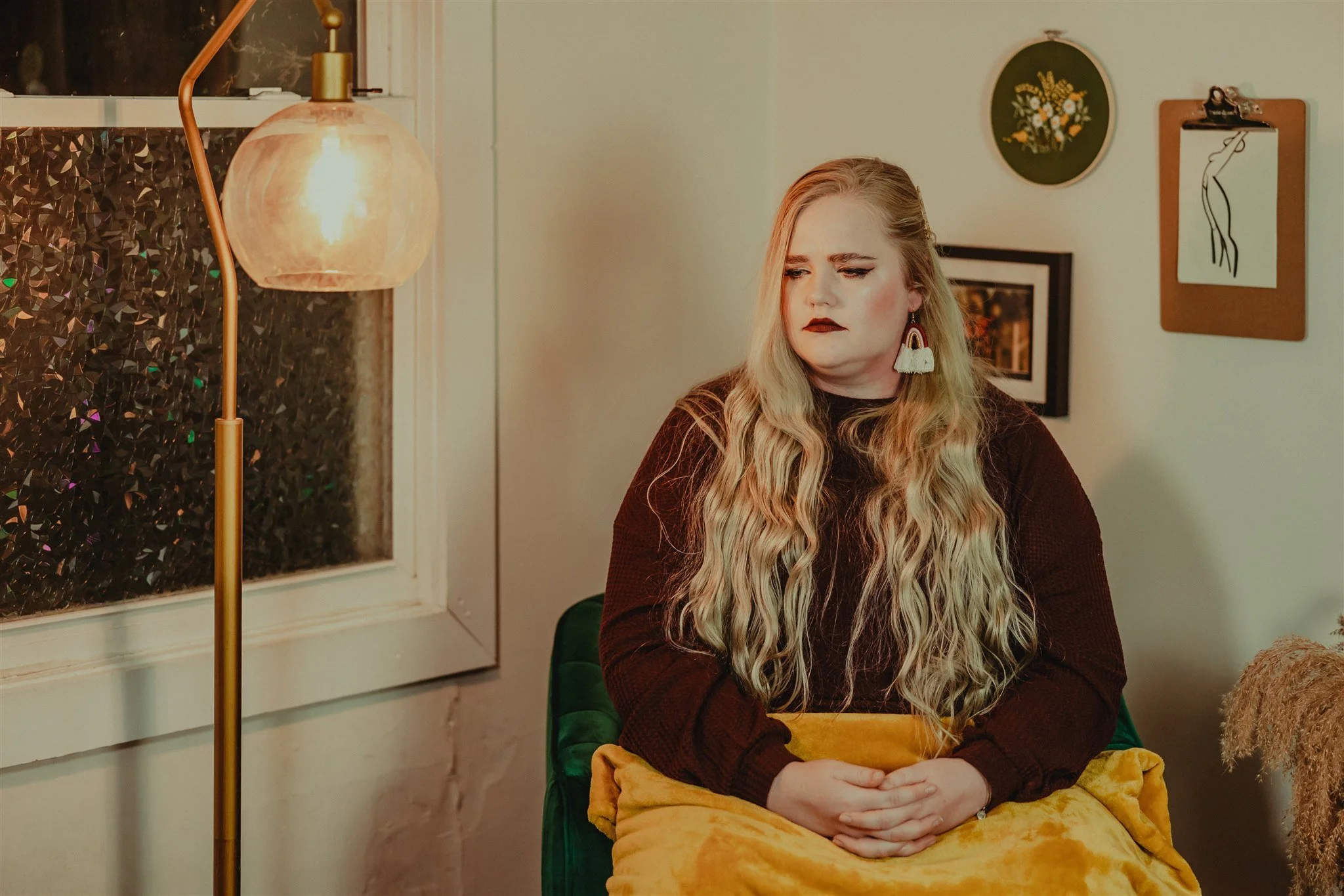 A woman with long blonde wavy hair, wearing dark lipstick, a dark top, and large earrings, sitting on a green chair with a yellow blanket in her lap during a trauma informed photography session in Columbia, Missouri.