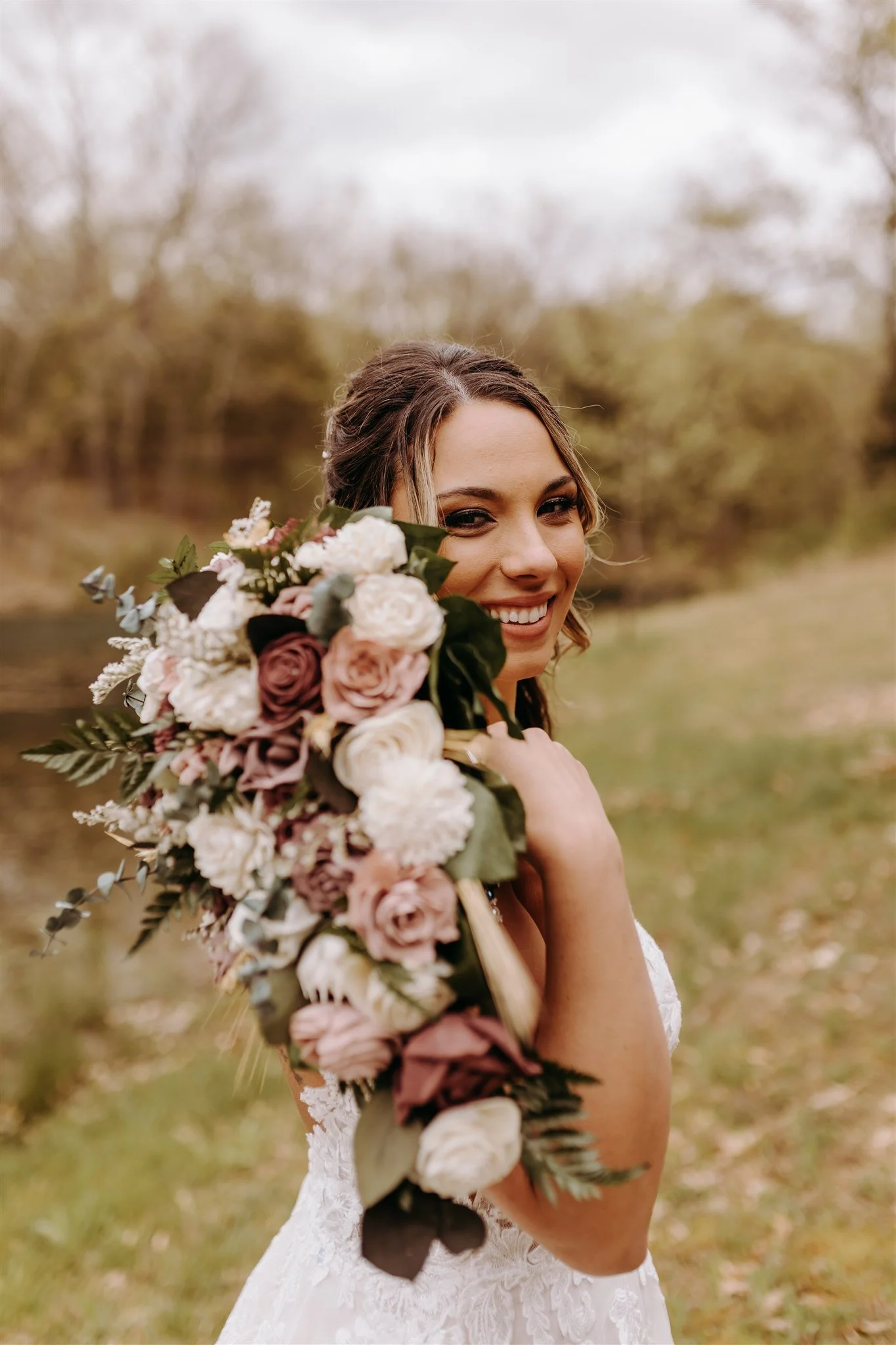 A bride holding a large bouquet of flowers outdoors, smiling at the camera with a blurred natural background at Rolla, MO rustic wedding venue, The Village. Photos by Columbia, MO Photographer Liv Strange Photography.