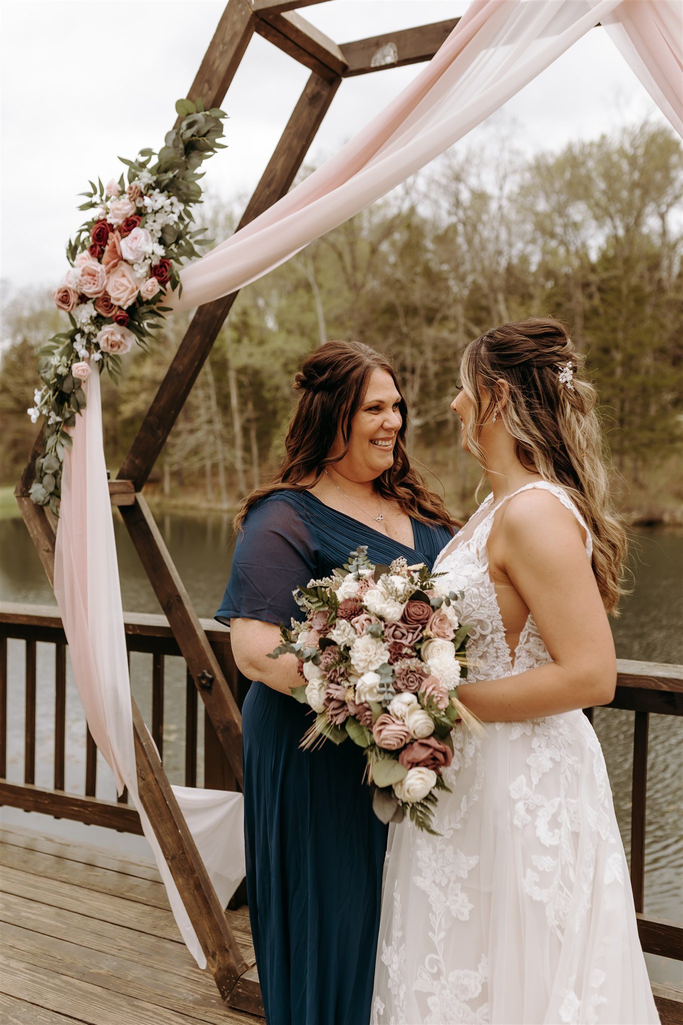 A bride and her mother, sharing an emotional moment during a wedding ceremony on a wooden deck by a lake at Rolla, MO rustic wedding venue, The Village. Photos by Columbia, MO Photographer Liv Strange Photography.