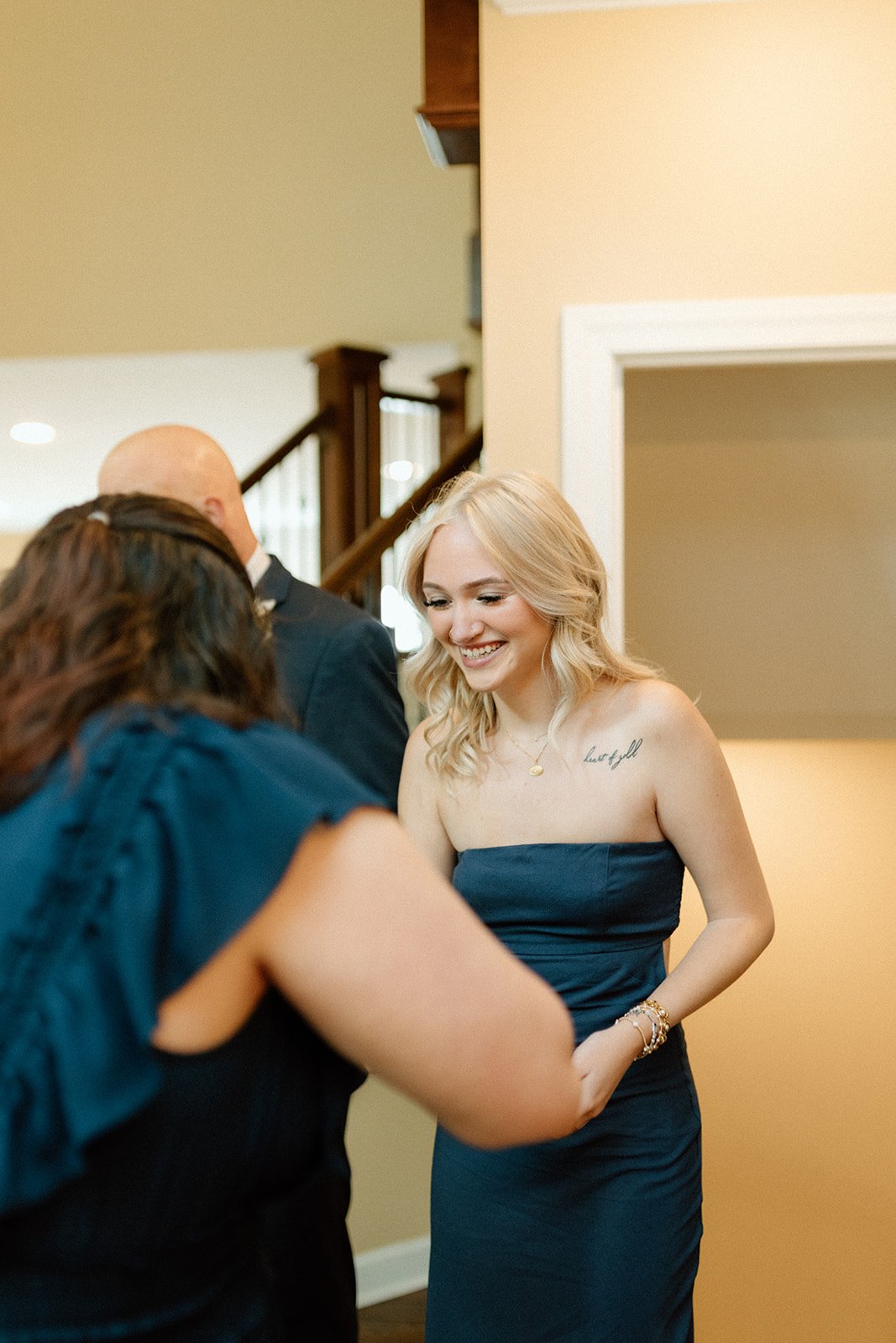 A woman with blonde hair in a strapless navy dress smiling as she interacts with others at a gathering indoors at an intimate backyard wedding in Columbia, MO.