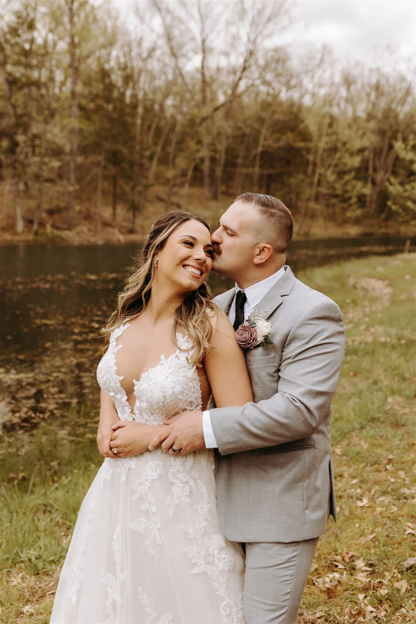 A bride and groom embracing outdoors next to a river, with trees in the background, during their wedding at Rolla, MO rustic wedding venue, The Village. Photos by Columbia, MO Photographer Liv Strange Photography.
