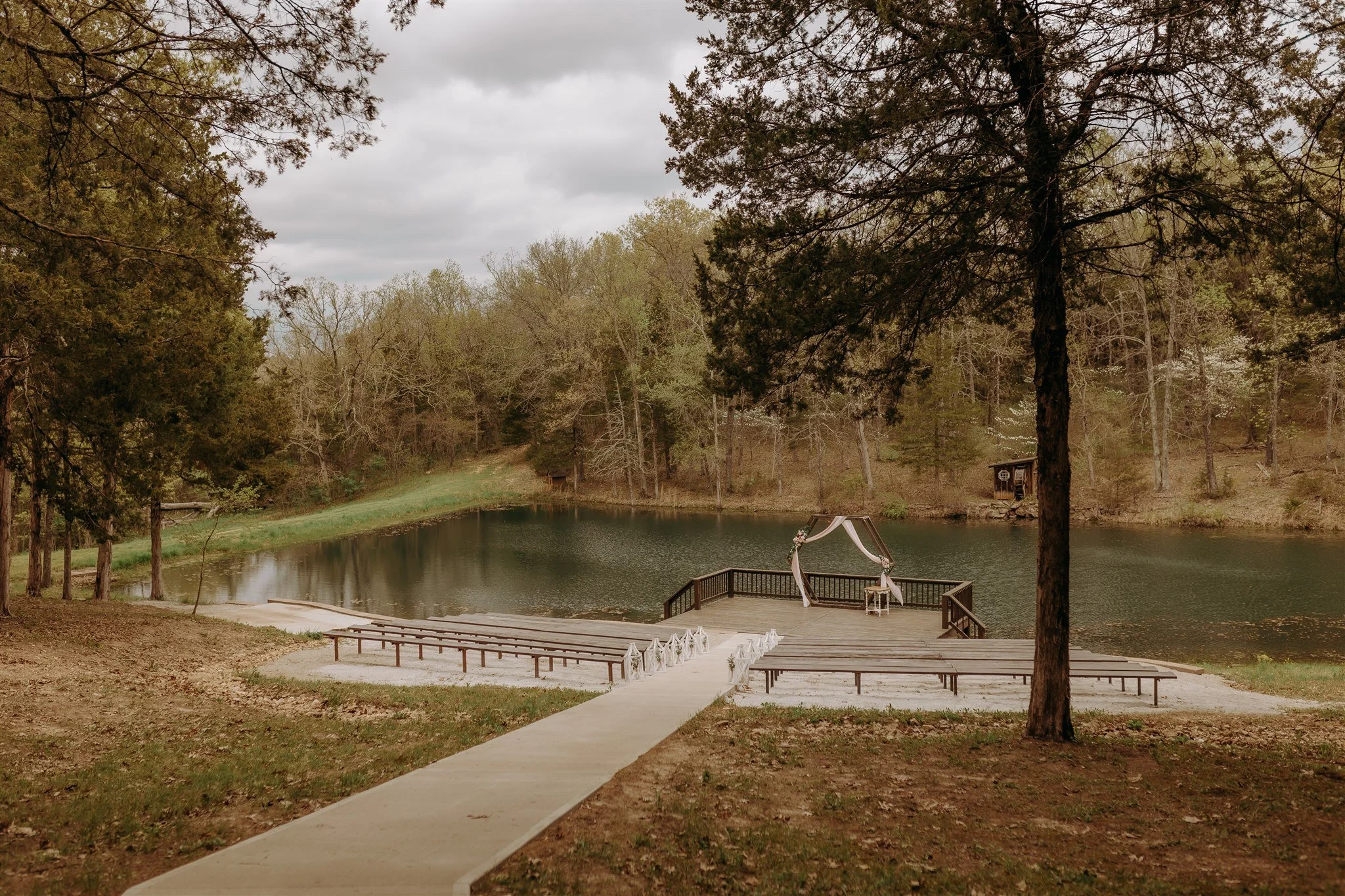 Wedding altar on a dock by a lake with seating, trees surrounding, and cloudy sky at Rolla, MO rustic wedding venue, The Village.