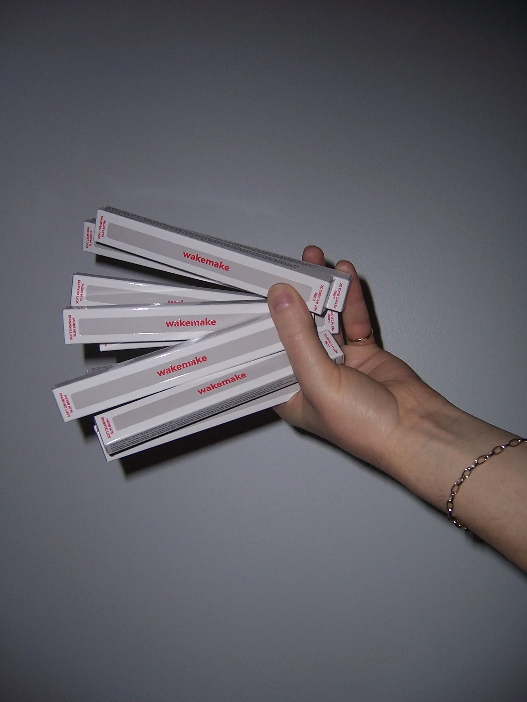 Hand wearing a gold bracelet hands a fistful of Wakemake branded eyebrow pencils in their boxes