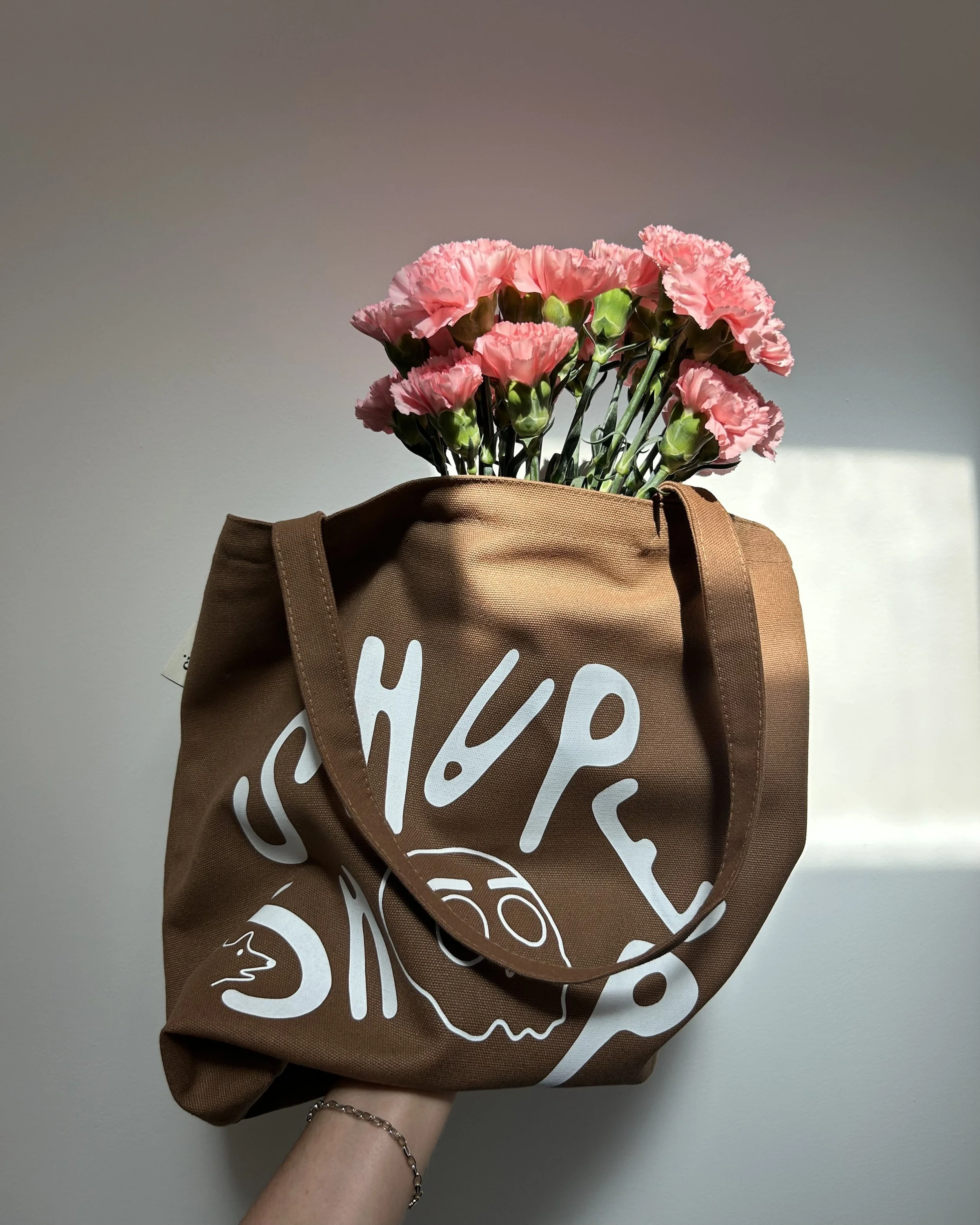 an arm wearing a gold bracelet holds up a brown tote bag filled with pink flowers that look like carnations. the bag has the letters "SHAPE SHOP" spelled out on the front. the "O" is a skull which is the brand's logo
