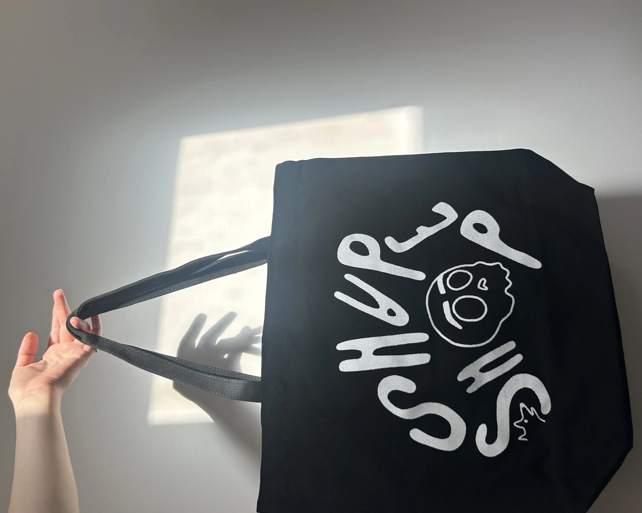a right hand comes up from the base of the photo and holds the handle of a black tote bag that reads "SHAPE SHOP". the image is flipped so it looks like the hand is holding the bag which is to the right of it.