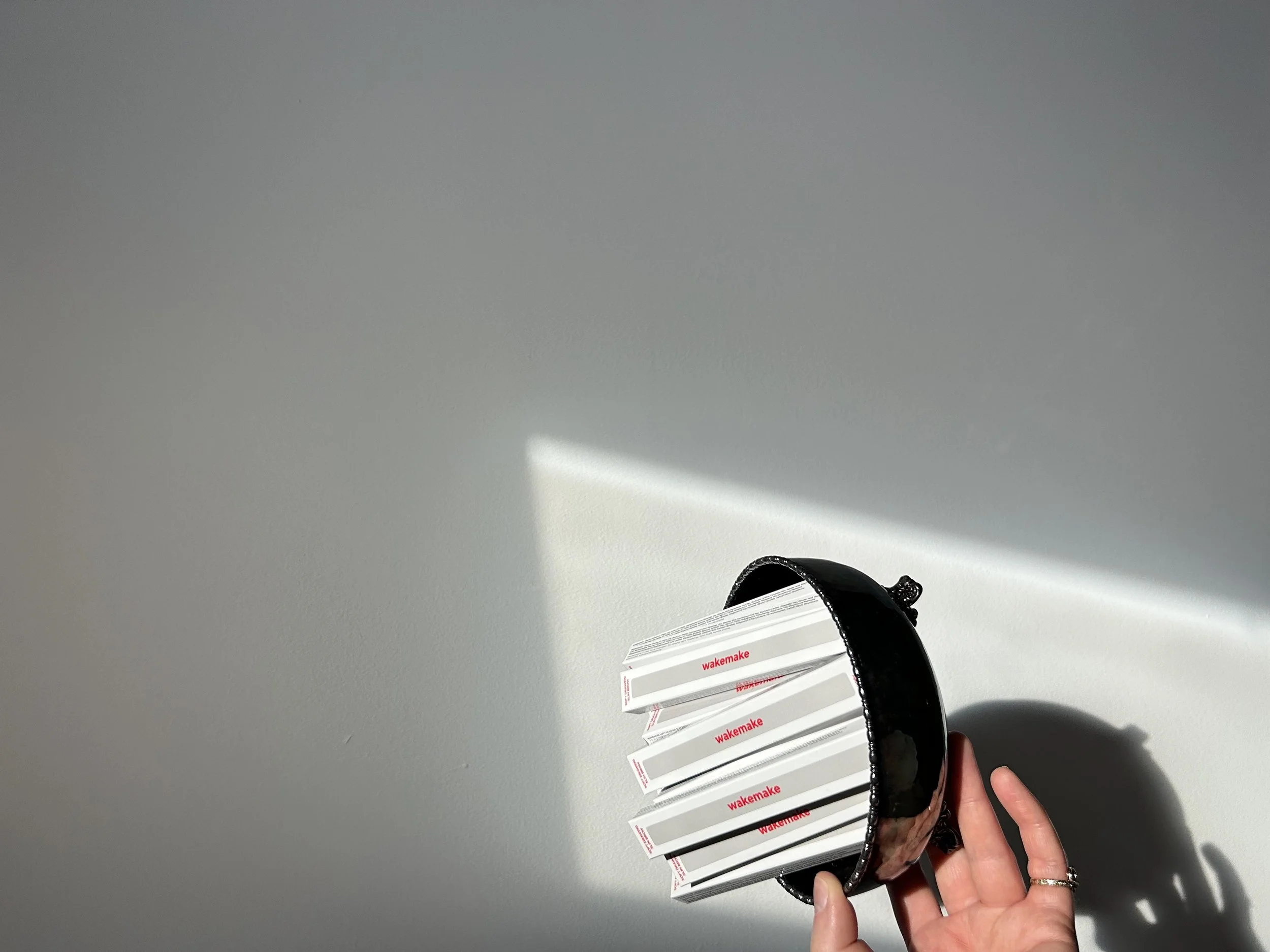 The fingers of a partially visible hand hold a bowl filled with eyebrow pencils still in their boxes. The hand and bowl are framed by a bright spot on the wall from a skylight above. The image is rotated so the bowl and its contents are tilted