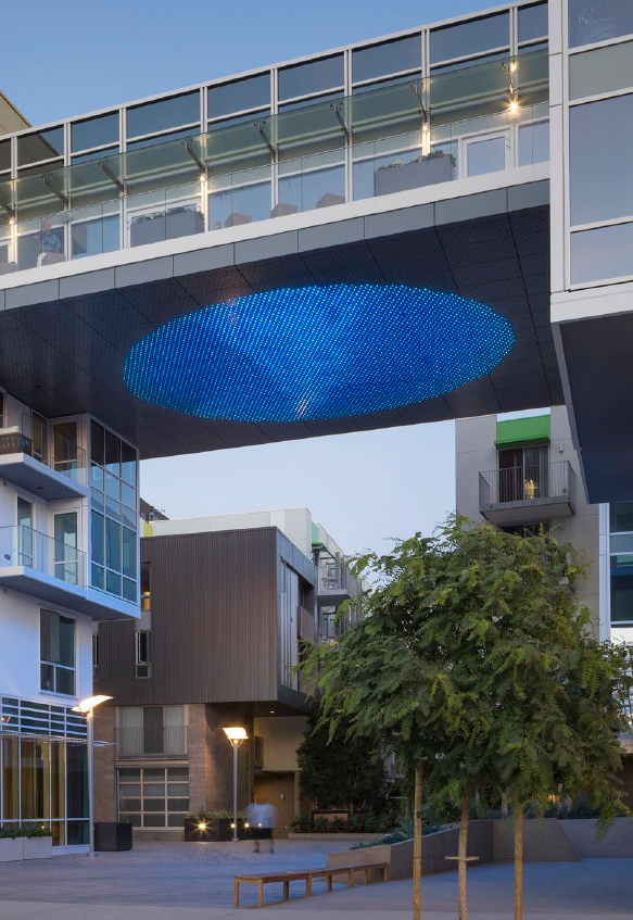 Modern urban courtyard with a futuristic building featuring an illuminated blue circular pattern under an overhanging structure, trees, benches, and surrounding contemporary architecture.