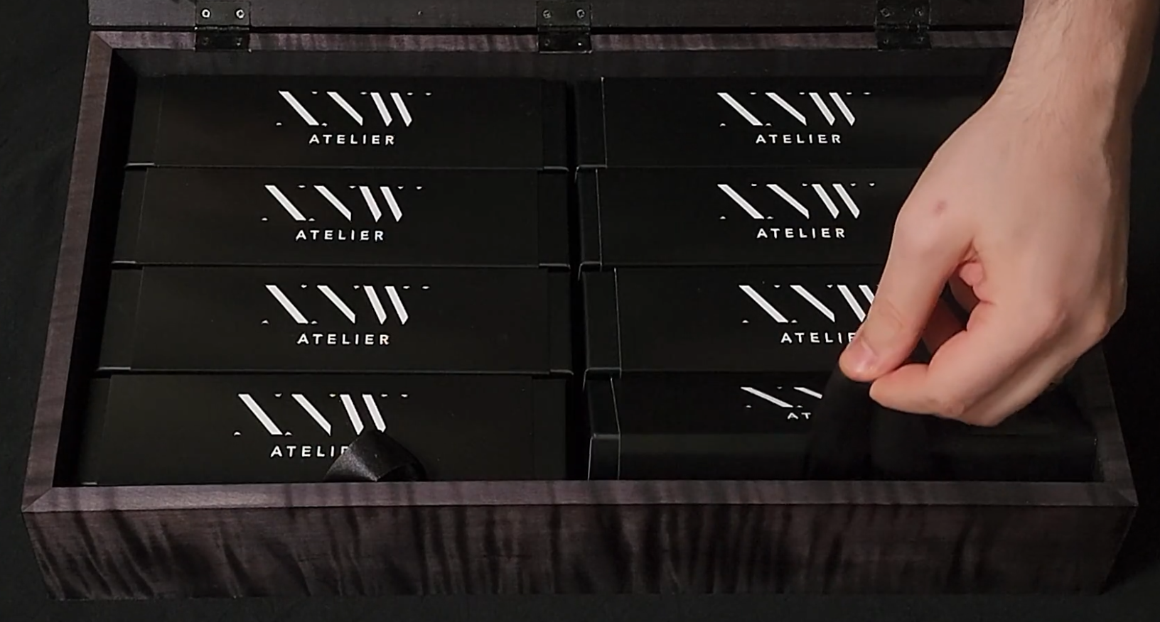 A hand reaching into a black wooden box with multiple black boxes labeled 'ATELIER' with white logo, arranged in a grid.
