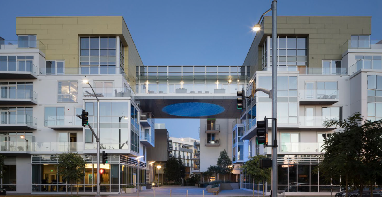 Modern apartment complex with interconnected building structures, glass balconies, and large windows, illuminated at dusk.
