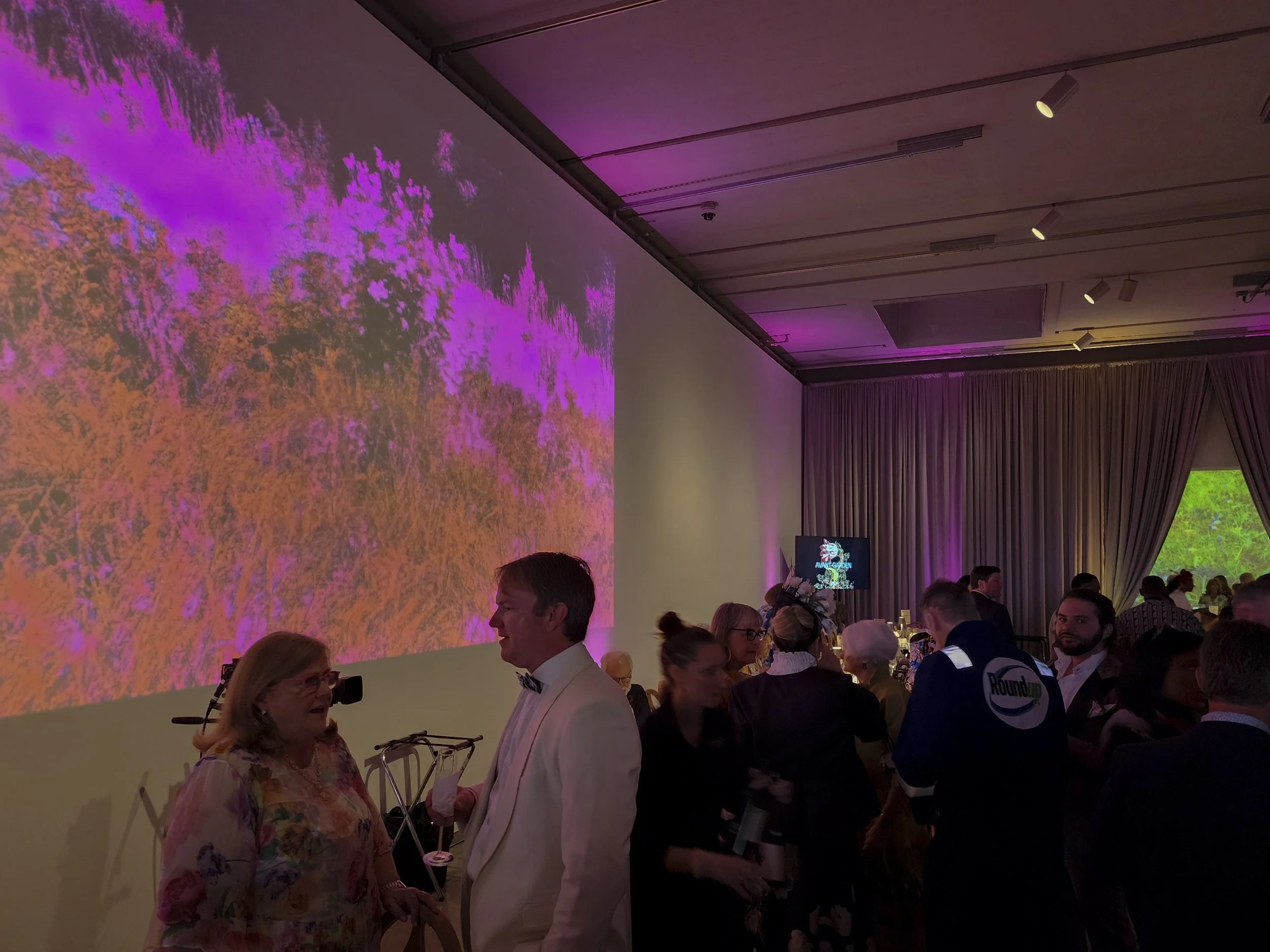 People attending an indoor event with a large colorful digital display on the wall, purple curtains, and some individuals wearing t-shirts with campaign logos.