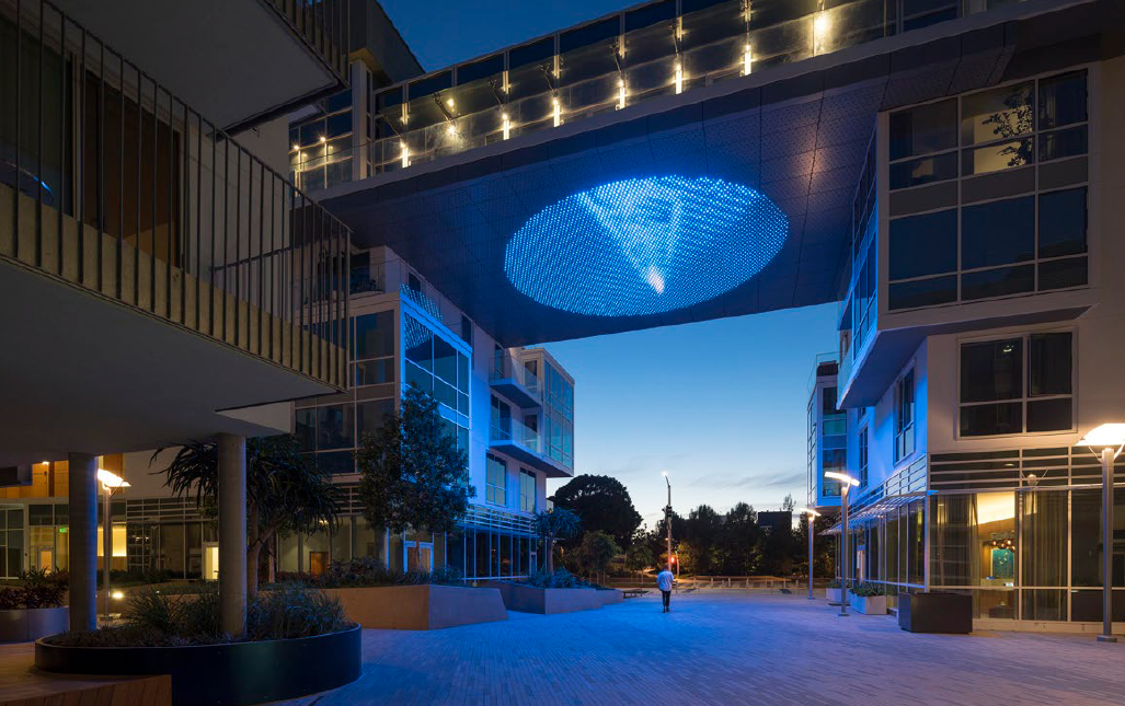 Nighttime view of a modern residential complex with illuminated blue lighting, including an overhead canopy with a spiral pattern and a person walking through the courtyard.