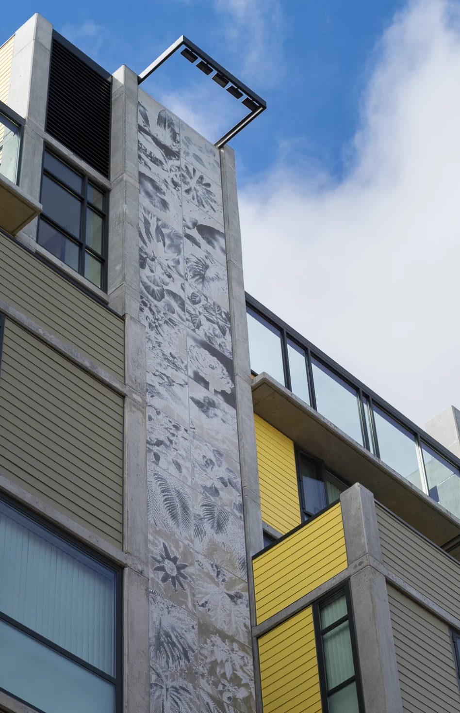 Close-up of a modern multi-story building with concrete walls, large glass windows, yellow balcony panels, and decorative tiles with black-and-white leaf patterns, against a blue sky with some white clouds.
