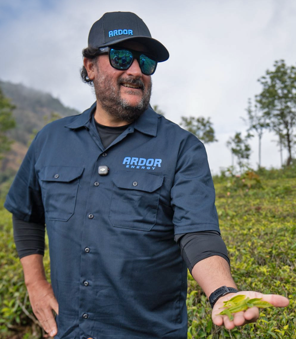 Man wearing sunglasses, a cap with 'ARDOR' logo, and a dark blue shirt with 'ARDOR ENERGY' emblem, smiling and holding a small green plant outdoors with trees and cloudy sky in the background.