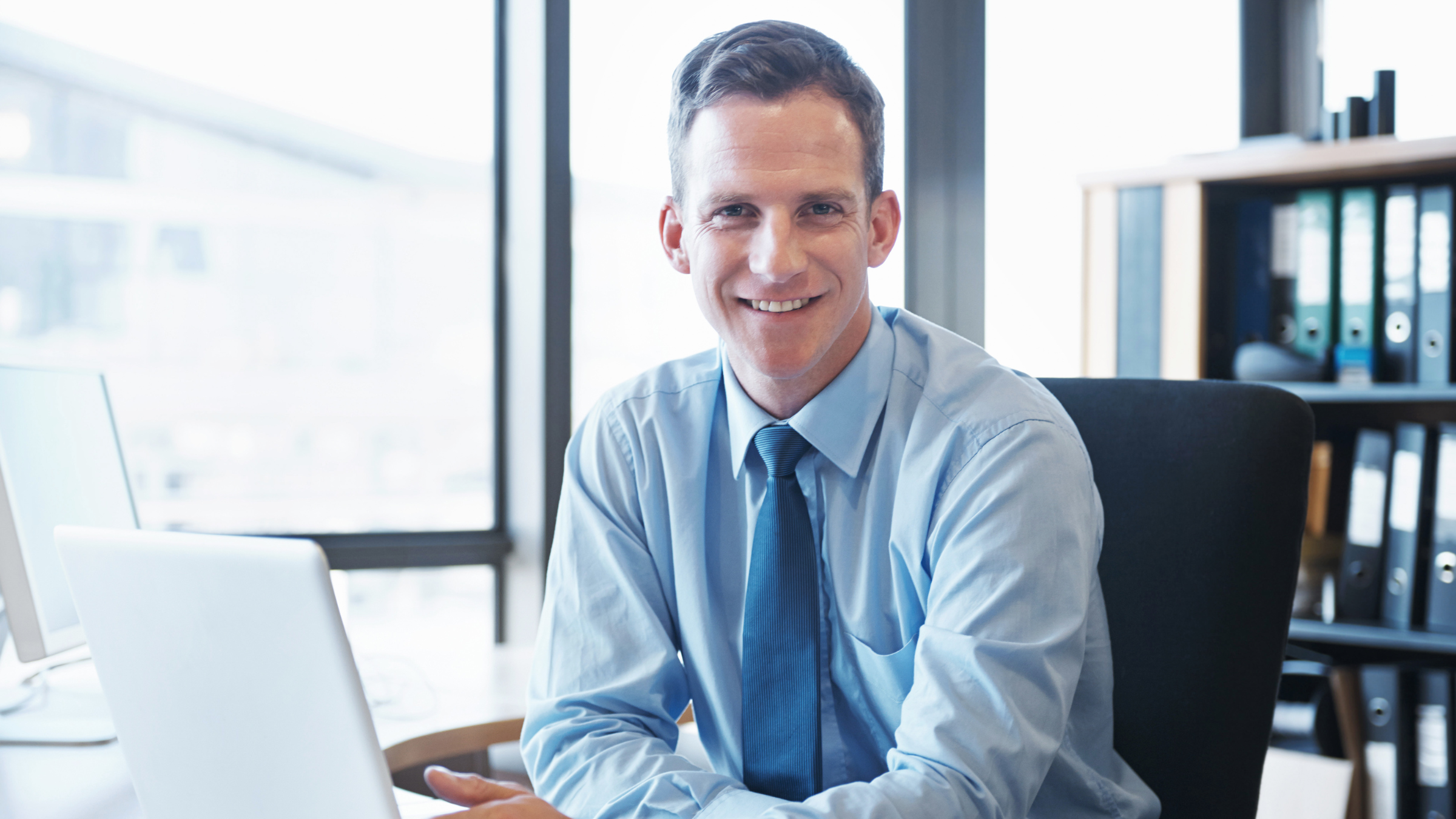A smiling man in business attire sitting at a desk in an office with large windows and shelves with binders.