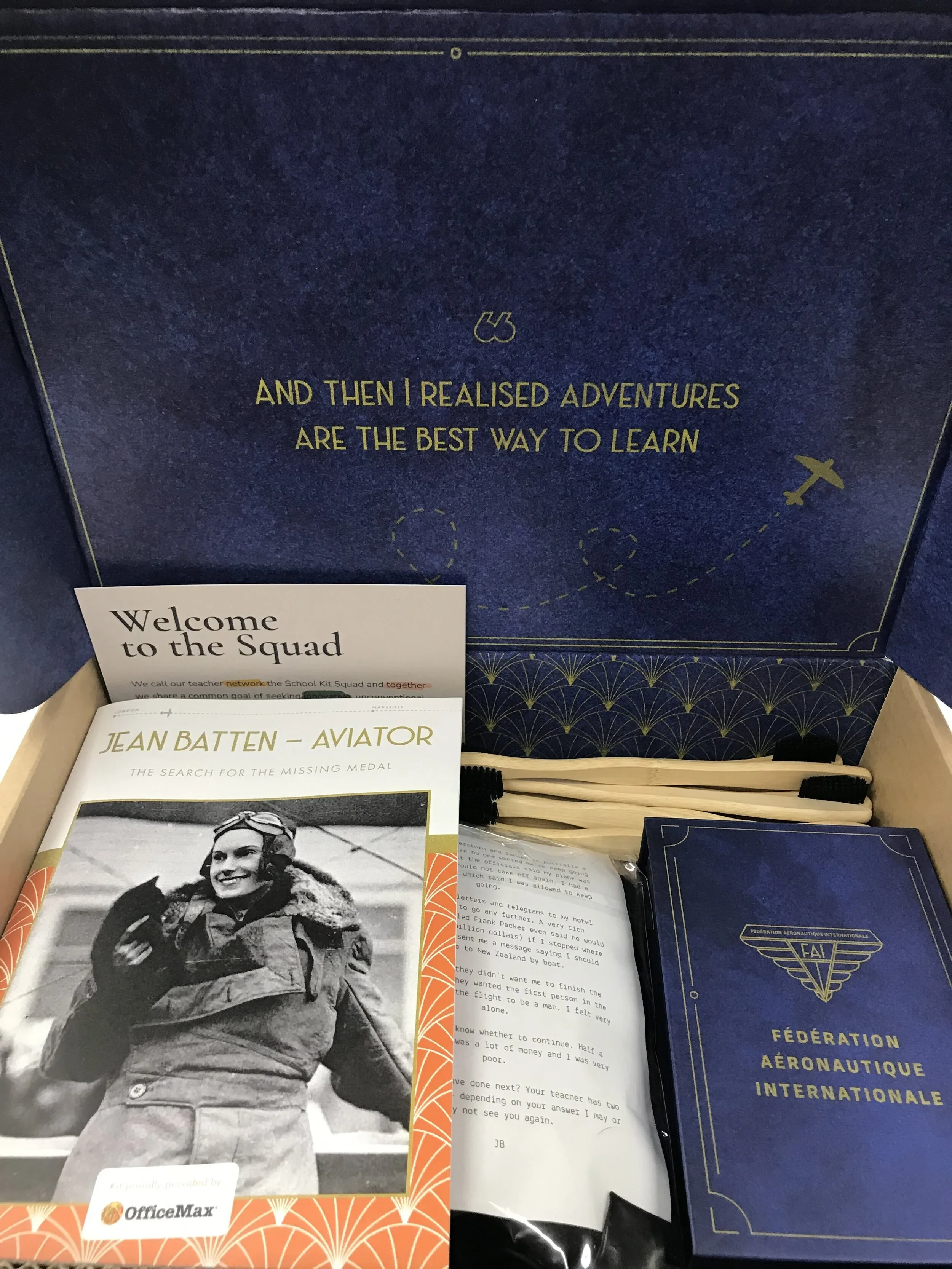 Open box containing aviation-themed educational materials, including a booklet titled 'Jean Batten - Aviator', a booklet from the Fédération Aéronautique Internationale, a welcome card, and some printed pages, with a blue interior lid that reads 'And then I realised adventures are the best way to learn'.