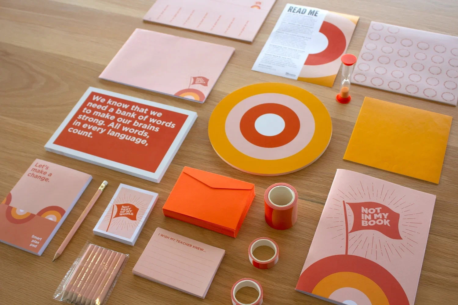 Arranged on a wooden table are various stationery items and posters featuring orange, pink, and white colors. Items include notebooks, a pencil, tape rolls, an hourglass, a stack of pink sticky notes, and posters with messages like 'We know we need a bank of words to make our brains strong' and 'Not in my book,' along with circular design elements.