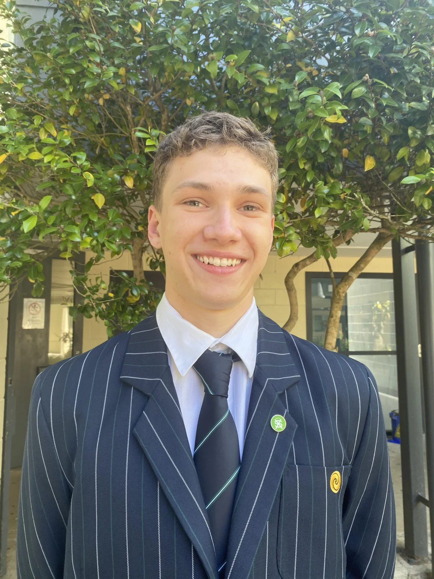 A young man with short, curly brown hair, smiling, wearing a navy blue pinstripe suit, white dress shirt, and a navy blue tie with green and white diagonal stripes, standing outdoors in front of a leafy green tree and a building.