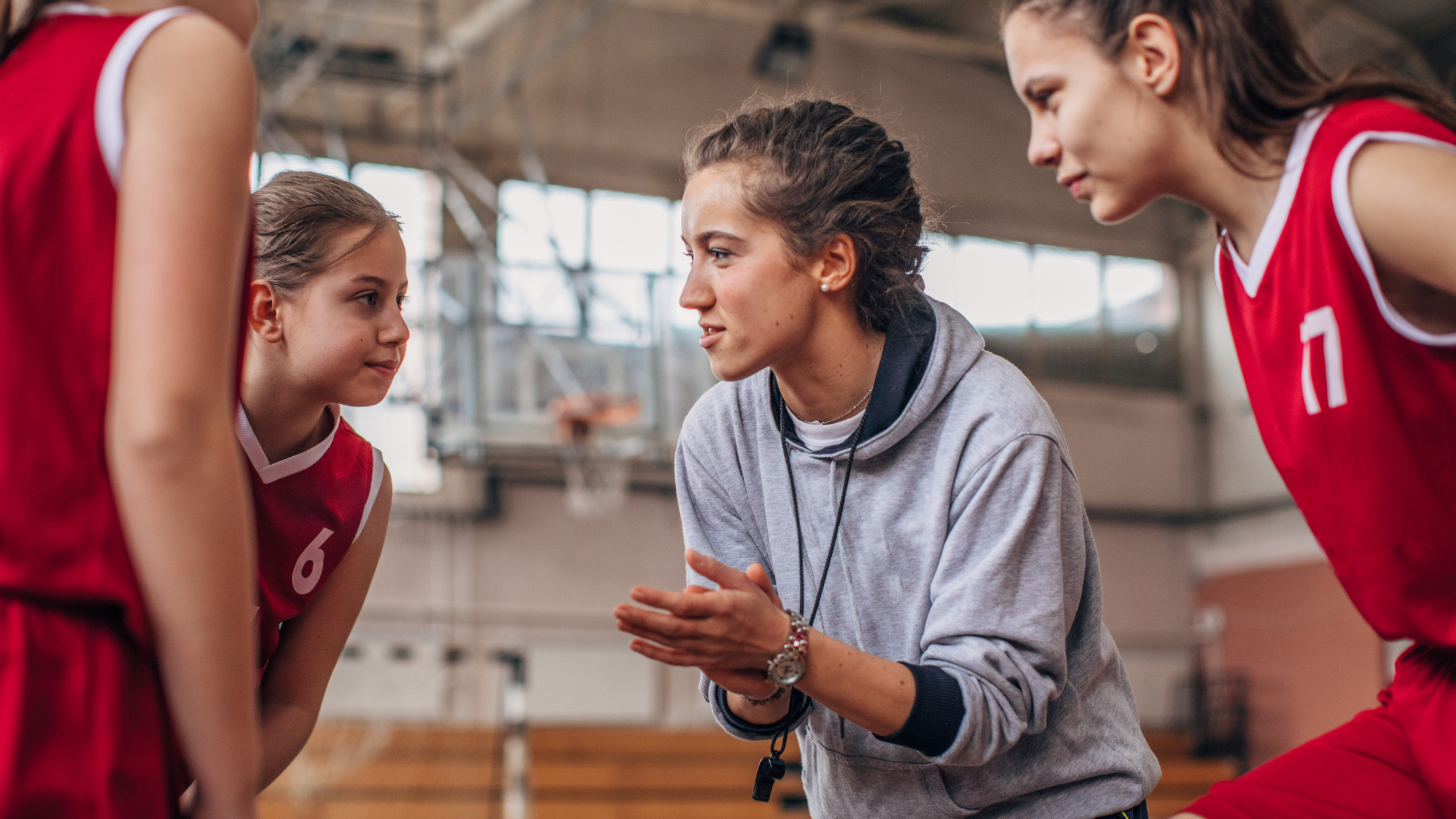 A female basketball coach in gray hoodie giving instructions to young female basketball players in red uniforms during a timeout in a gymnasium.