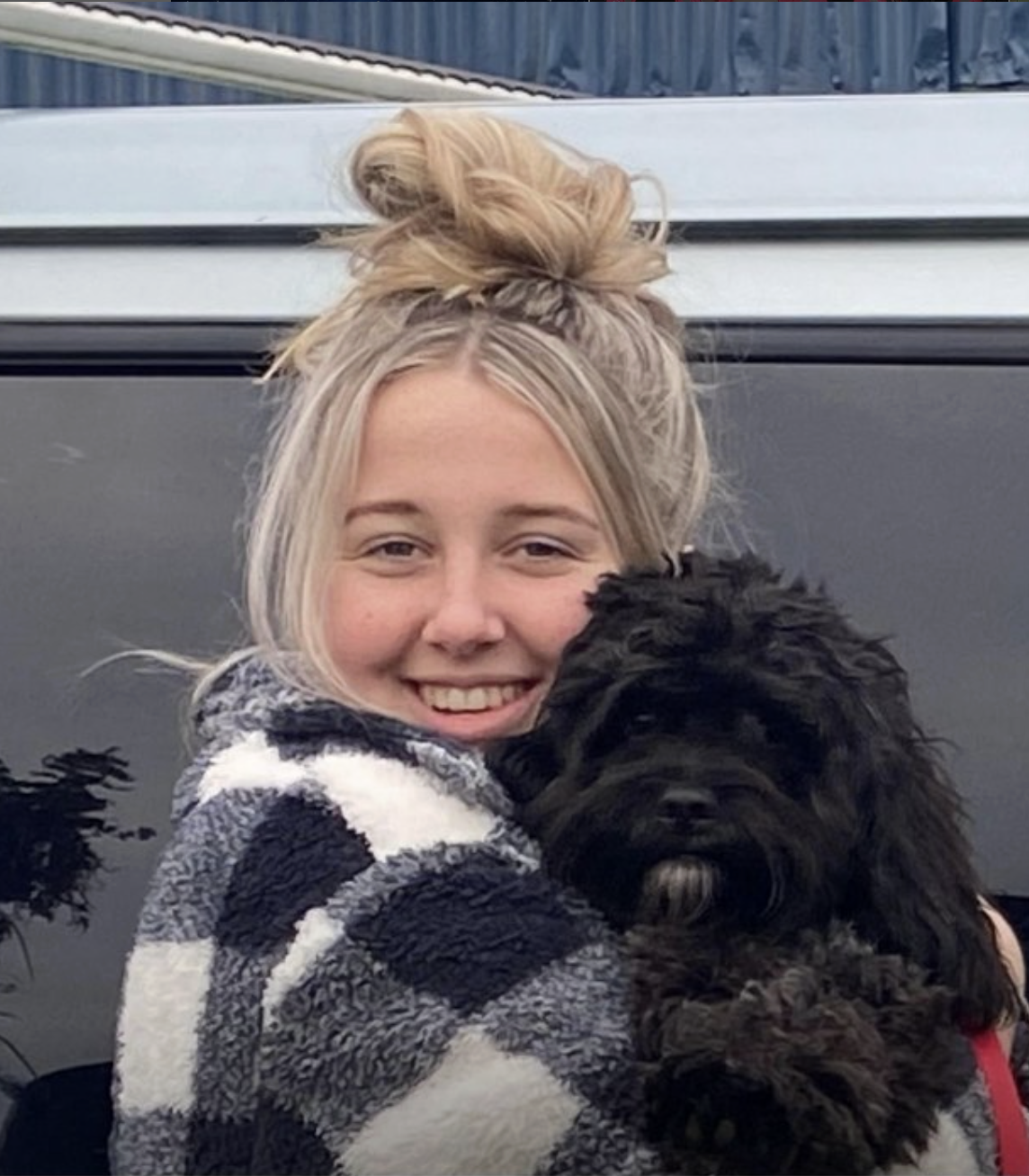 A young woman with blonde hair in a messy bun is smiling while hugging a black fluffy puppy outside near a car.