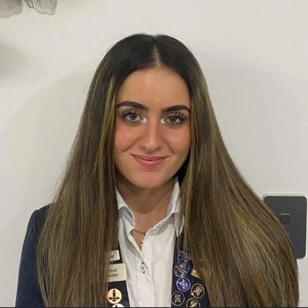 A woman with long brown hair, wearing a white collared shirt and a dark blazer, standing against a white wall.