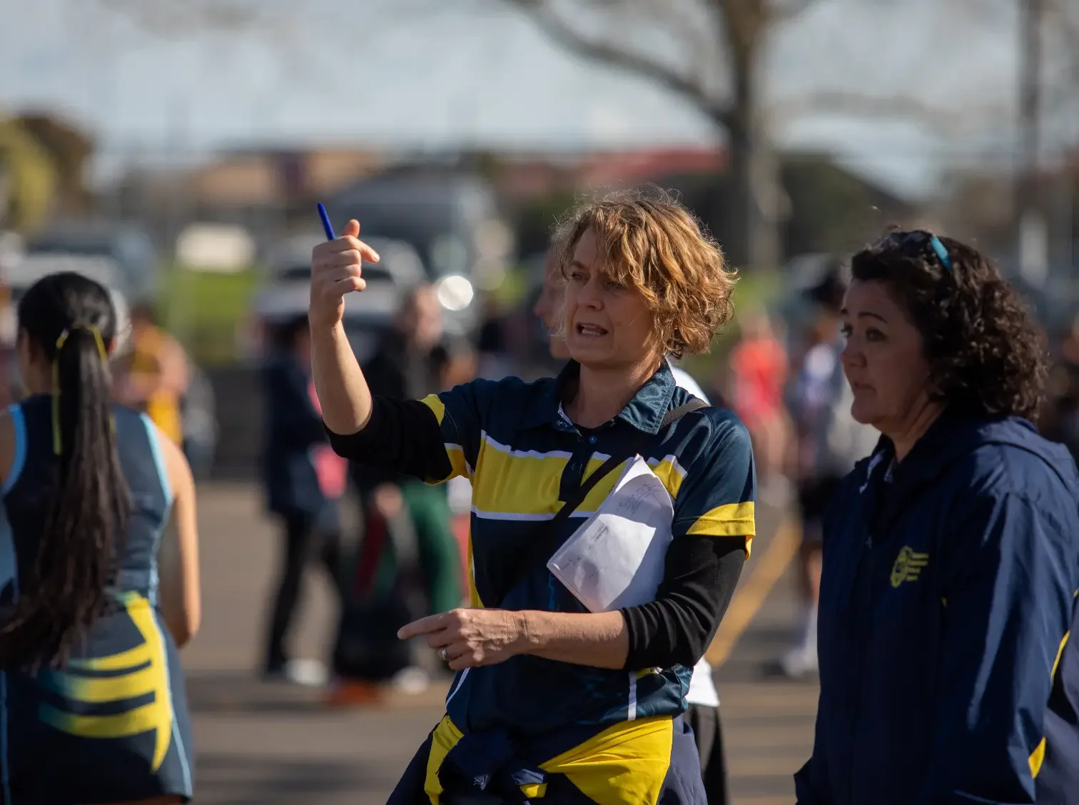 A woman with curly hair in a sports uniform holding a clipboard, giving instructions during a sports event, with other athletes nearby.