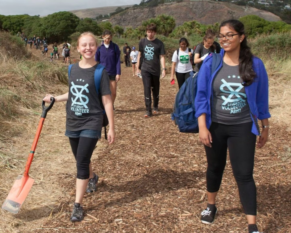 Group of young volunteers participating in a cleanup walk on a dirt trail in a natural outdoor setting with trees and hills in the background. They are wearing matching volunteer shirts and carrying backpacks, with one girl holding a trash picker.