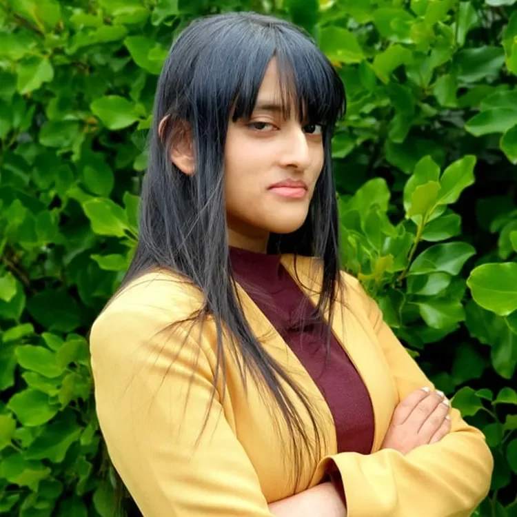 A woman with long dark hair standing outdoors in front of green leaves, wearing a yellow blazer and a maroon top, crossing her arms.