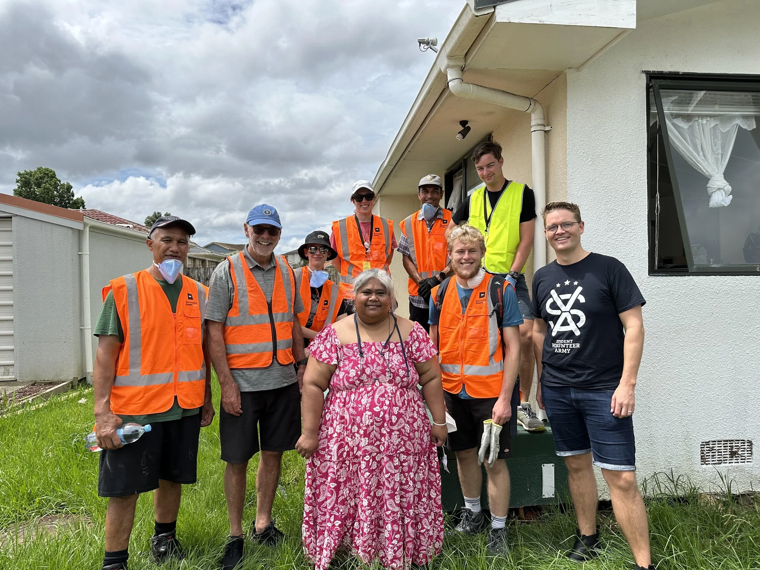 Group of volunteers, some wearing orange safety vests, standing outside a house on a cloudy day. One woman is wearing a pink dress, and others are smiling at the camera. The house has a white exterior with a window and a small porch.