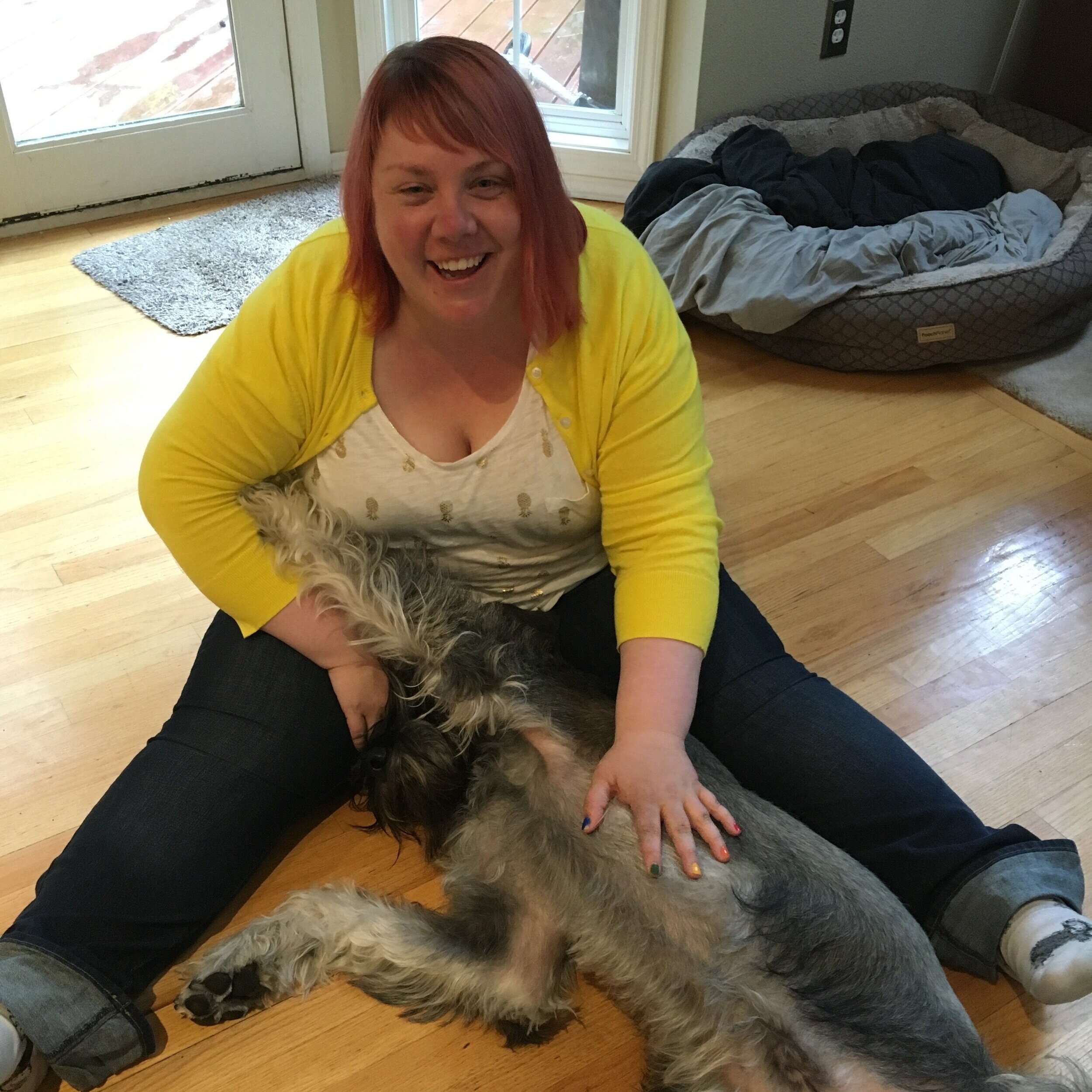 Manda sits on the floor smiling at the camera. In her lap is a big, gray dog that’s laying on his back.