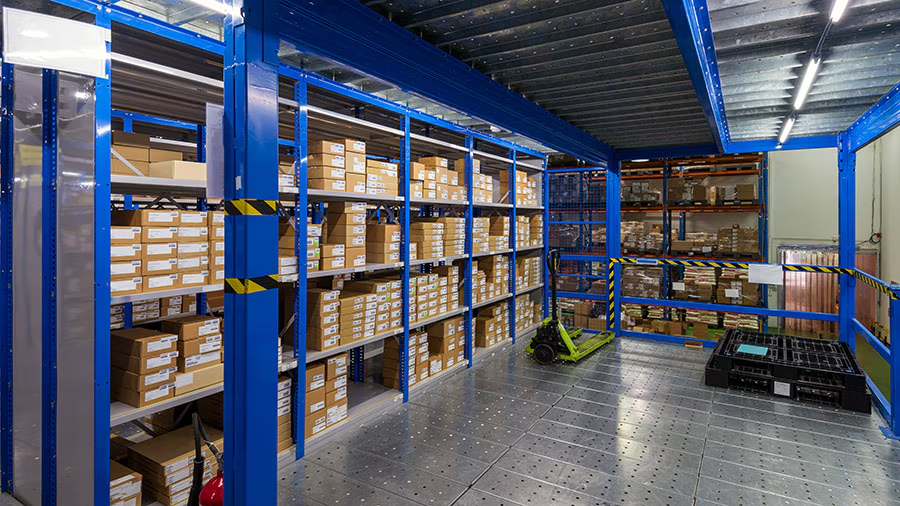 Industrial warehouse with blue shelving units filled with cardboard boxes, a green pallet jack, and black pallets on a gray metal floor, separated by caution tape.