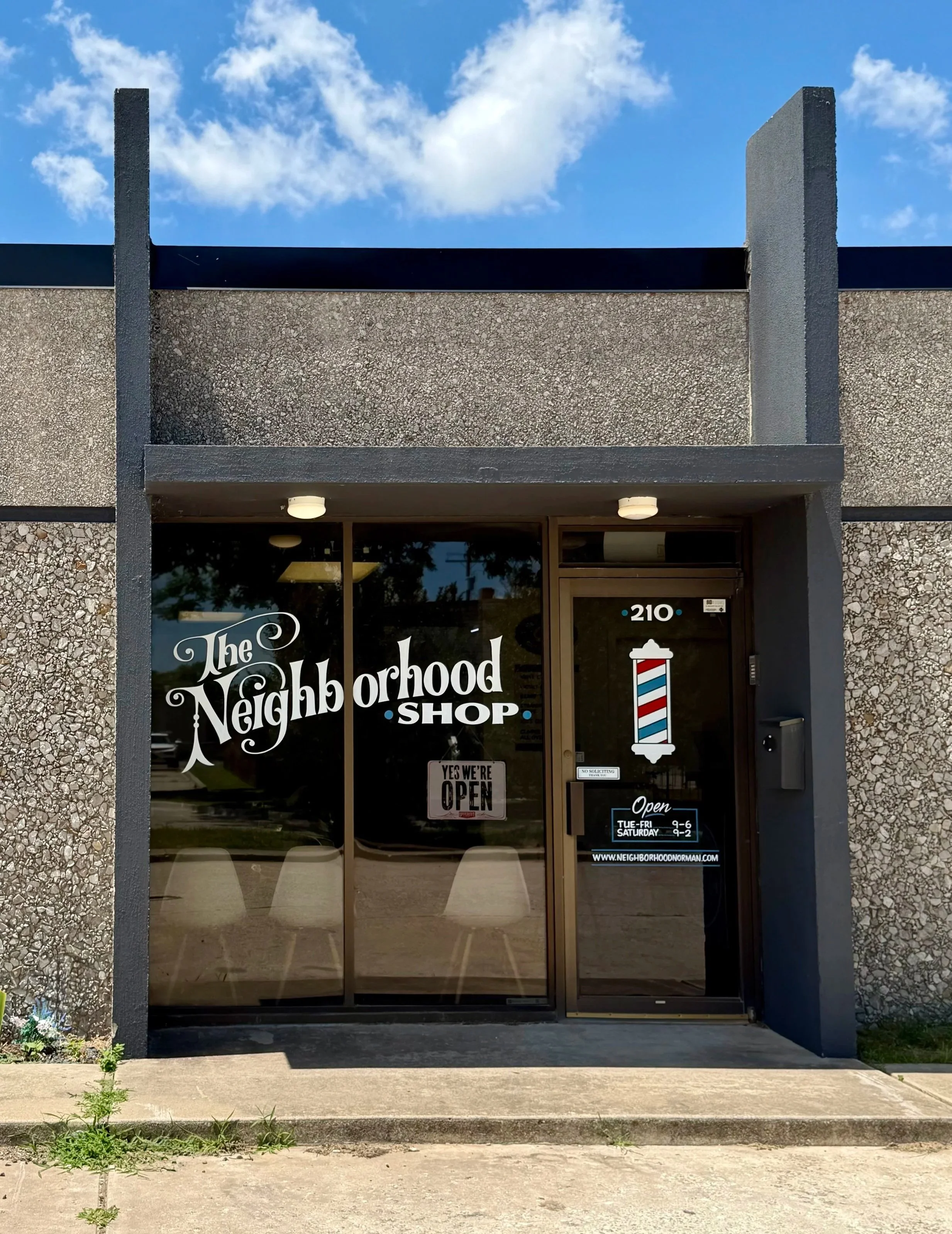 The clean storefront of The Neighborhood Shop with glass doors, displaying the business name, hours, website, and a barber pole graphic.