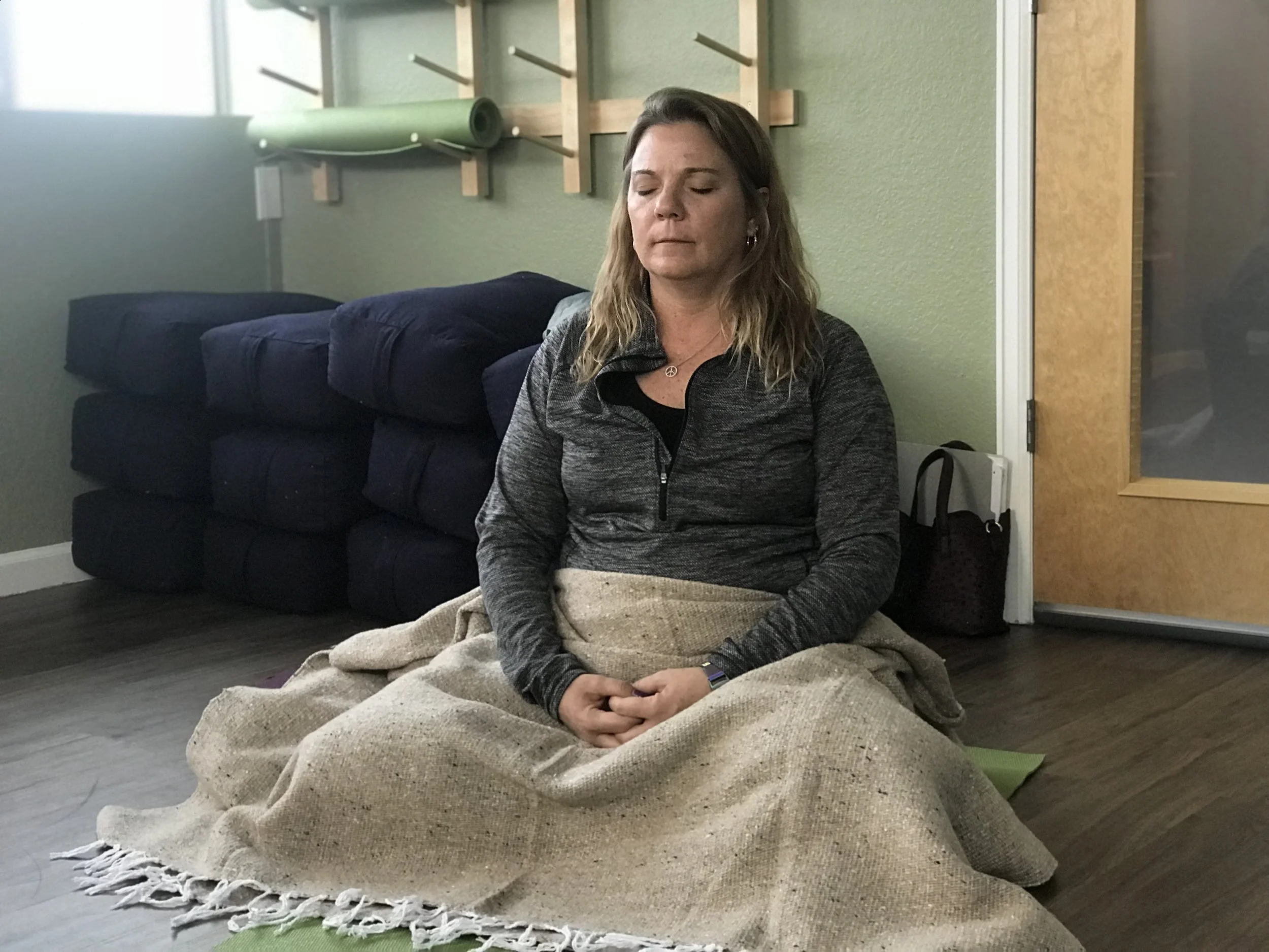 A woman with shoulder-length brown hair, wearing earrings and a black and gray athletic jacket, is sitting cross-legged on the floor with her eyes closed. She is covered with a beige blanket, and behind her is a dark blue couch with pillows. She appears to be meditating or resting in a room with light green walls and wooden decor.